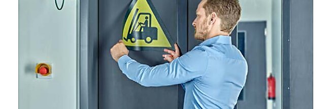 A man in a blue shirt is affixing a triangular caution sign with a forklift symbol to a gray door using tesa tape. A red emergency button and green exit sign are visible on the left wall, and a fire extinguisher is in the background. (This text has been generated by AI)