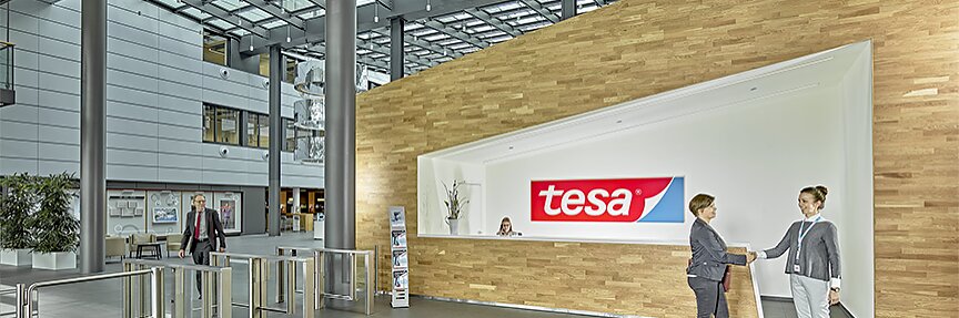 In a modern office lobby with a glass ceiling and a wooden reception desk displaying a tesa sign, two people are shaking hands in front of the desk, while another person is visible walking near turnstiles. Plants and wall displays enhance the decor. (This text has been generated by AI)