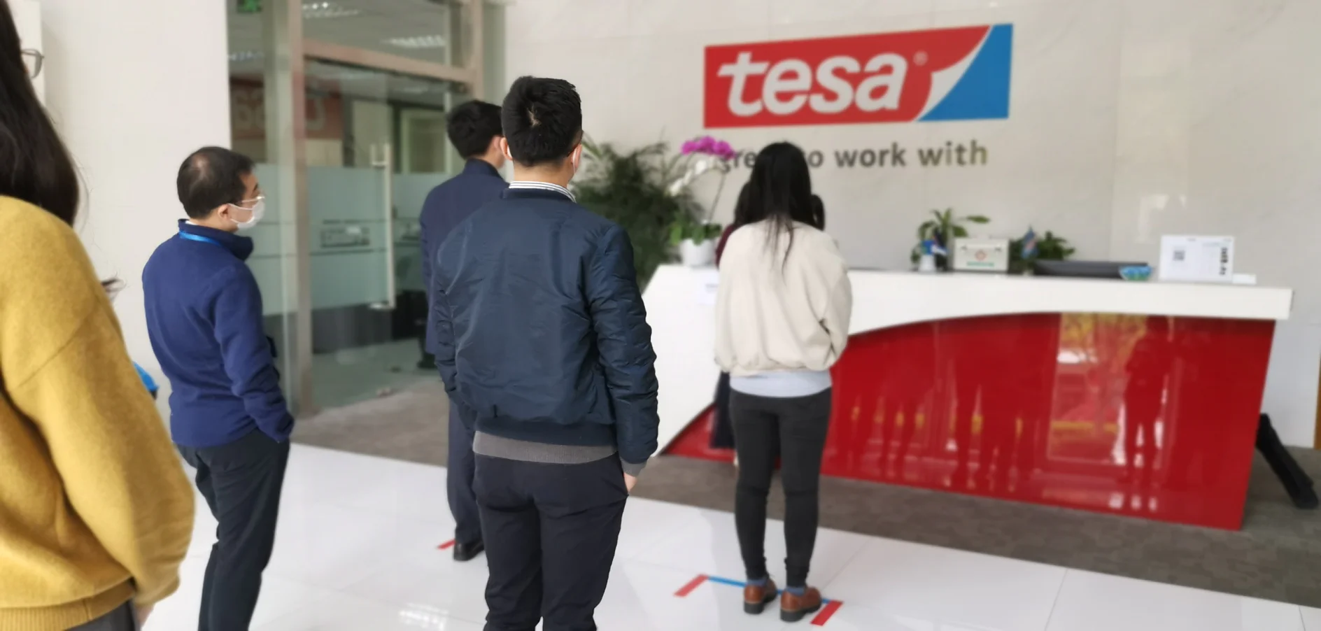 A group of people stand in line facing a reception desk inside a building with a sign reading tesa. The floor is white, and the desk is red with a white top. Some individuals are wearing jackets and someone is wearing a mask. (This text has been generated by AI)