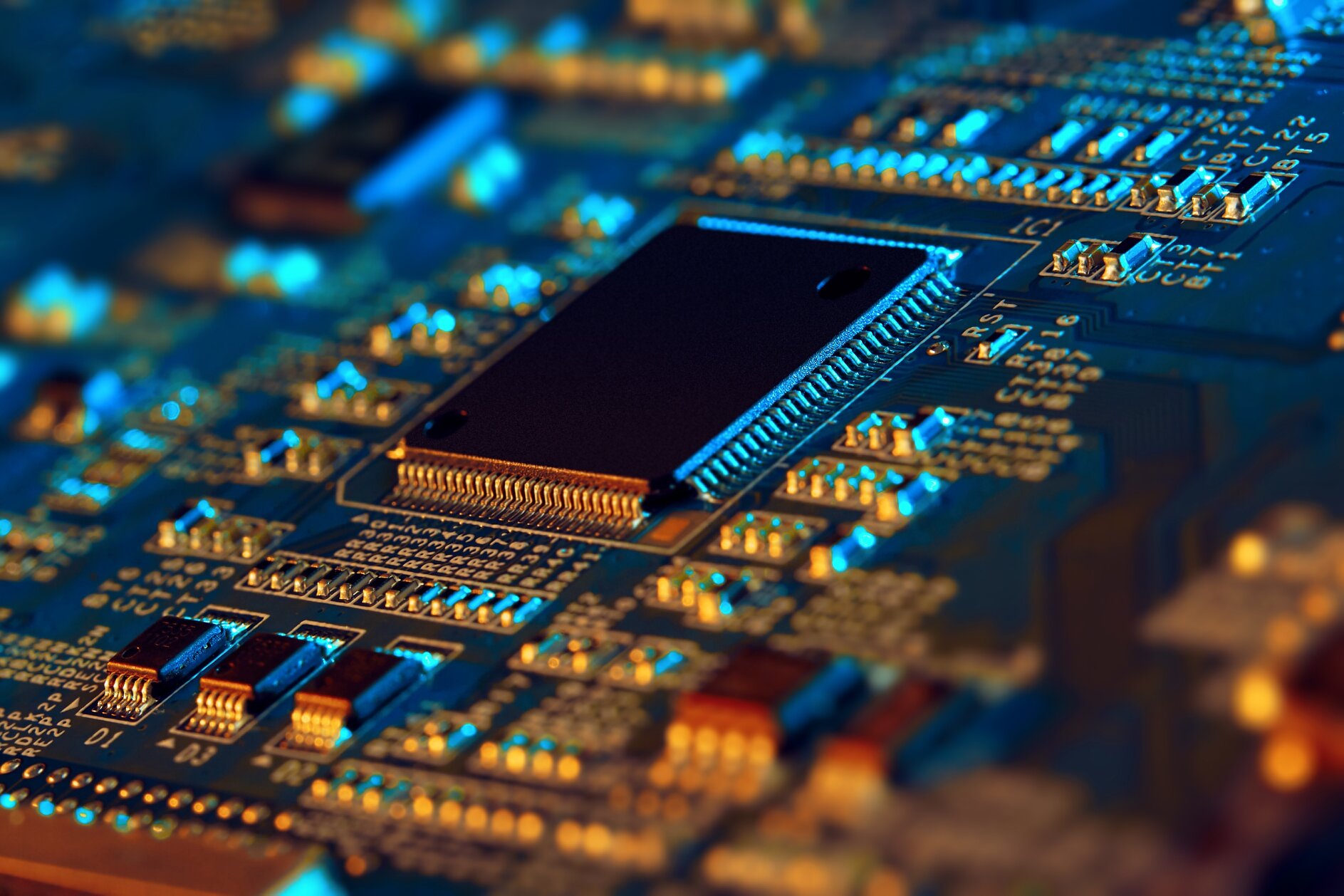 Close-up of a computer circuit board with electronic components. A central black microchip is prominently featured, surrounded by various smaller components like resistors and capacitors, with blue and orange lighting reflecting off the surface. (This text has been generated by AI)