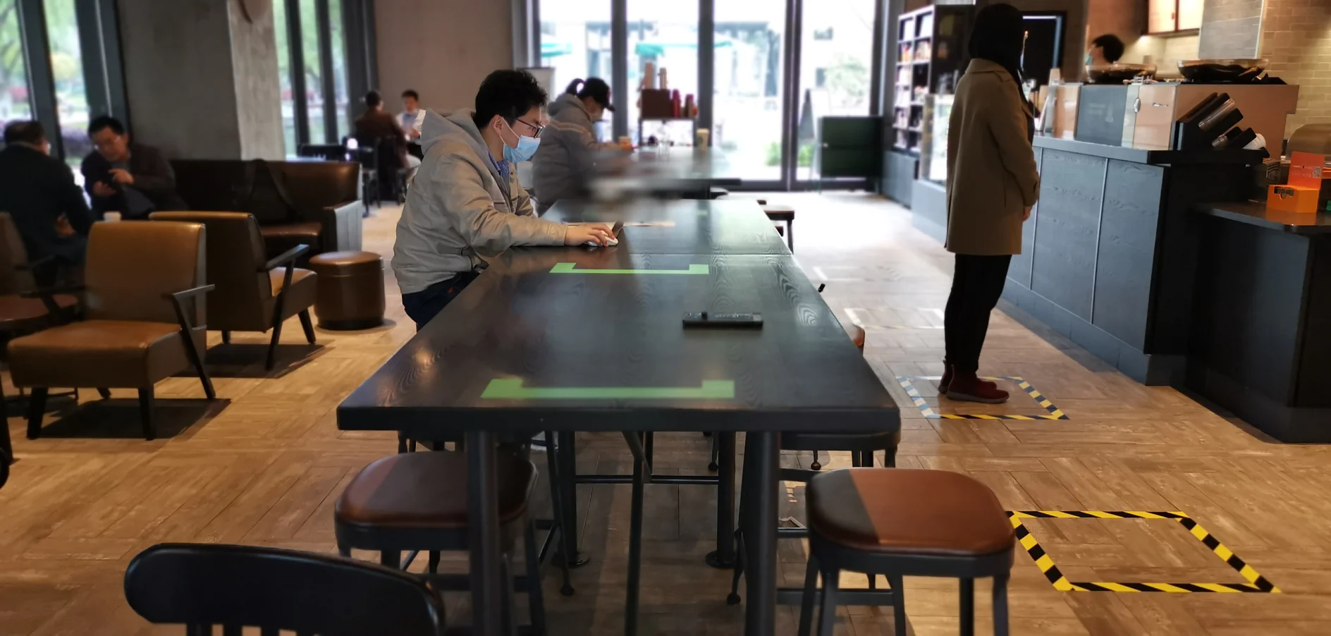 A cafe interior with several people seated at tables. Two individuals are sitting at a large table, one wearing a mask and using a smartphone. A woman stands at the counter. Social distancing markers made of tesa tape are visible on the floor. (This text has been generated by AI)