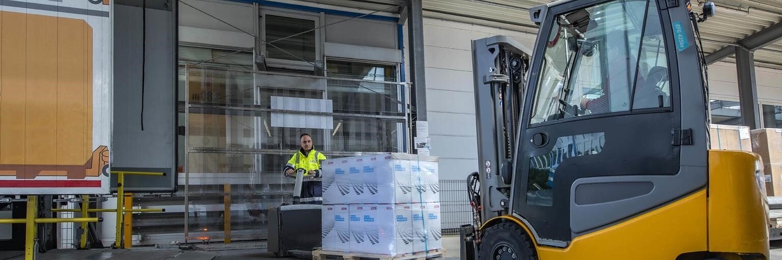 A forklift transports a pallet of goods from a warehouse, secured with tesa tape. A person in a high-visibility jacket operates the forklift. The area is covered with a metal roof, and a loading dock with a partially open door is visible in the background. (This text has been generated by AI)