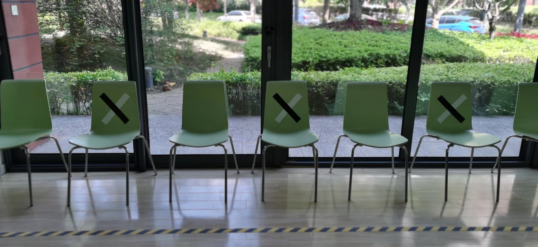 Six plastic chairs are lined up in front of large windows. Three chairs have black X marks on their backs, suggesting they are not for use. Outside, bushes and a path are visible. A floor marking tesa tape is on the ground. (This text has been generated by AI)