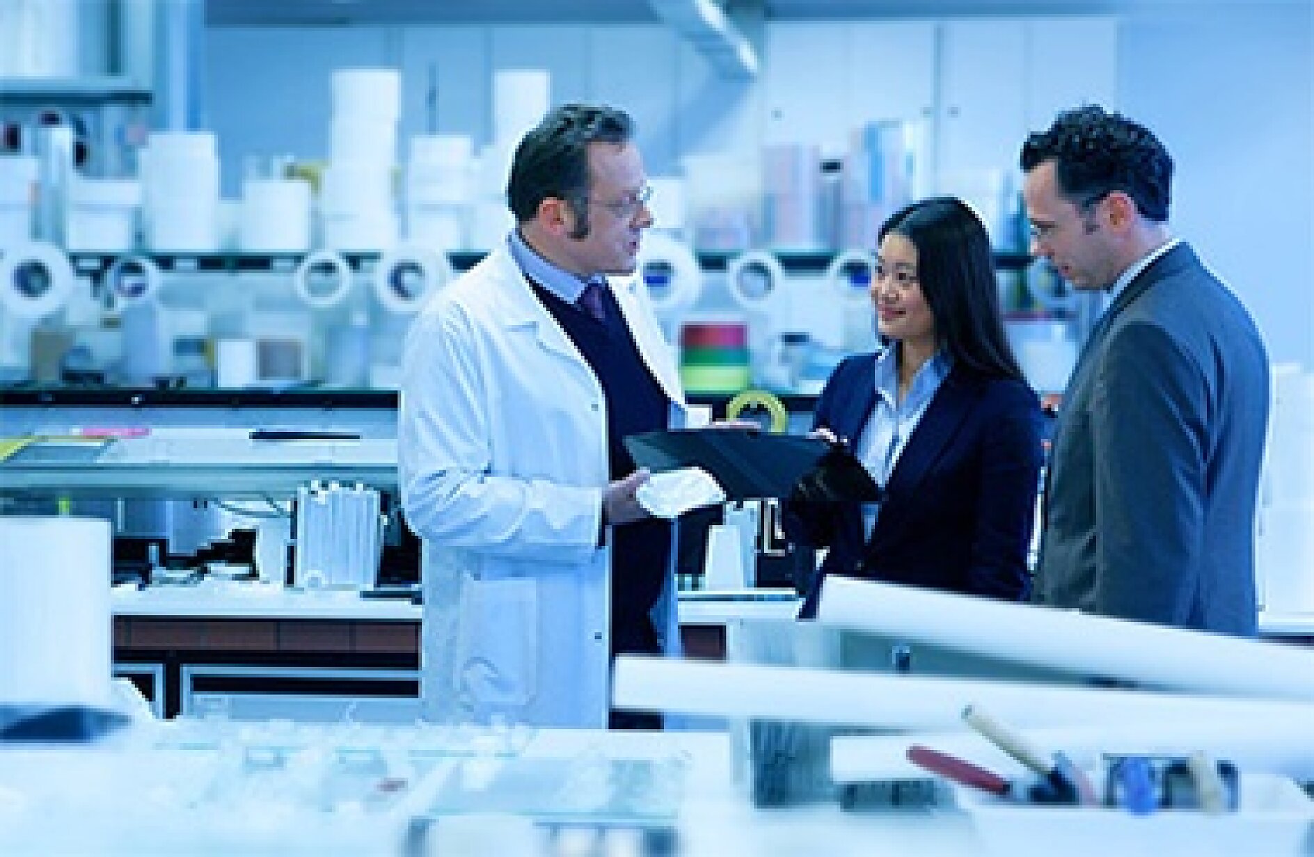 A man in a lab coat is demonstrating a product to a woman and a man in business attire within a laboratory environment. Shelves filled with scientific equipment and supplies can be seen in the background. The scene conveys a professional and focused atmosphere, with an emphasis on the usage of tesa tape. (This text has been generated by AI)