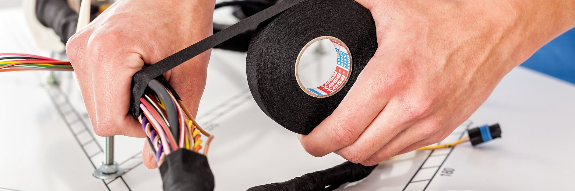 A person is wrapping a bundle of colorful wires with black tesa tape. The wires are laid out on a table, with additional tools and a spool of tesa tape visible. The background is a neutral, blurred setting. (This text has been generated by AI)