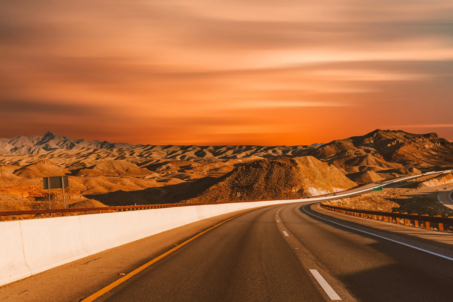 A deserted highway curves through a barren desert landscape at sunset. The sky is a deep orange with streaks of clouds, and distant rocky hills outline the horizon. A concrete barrier, affixed with tesa tape, runs along the road. (This text has been generated by AI)