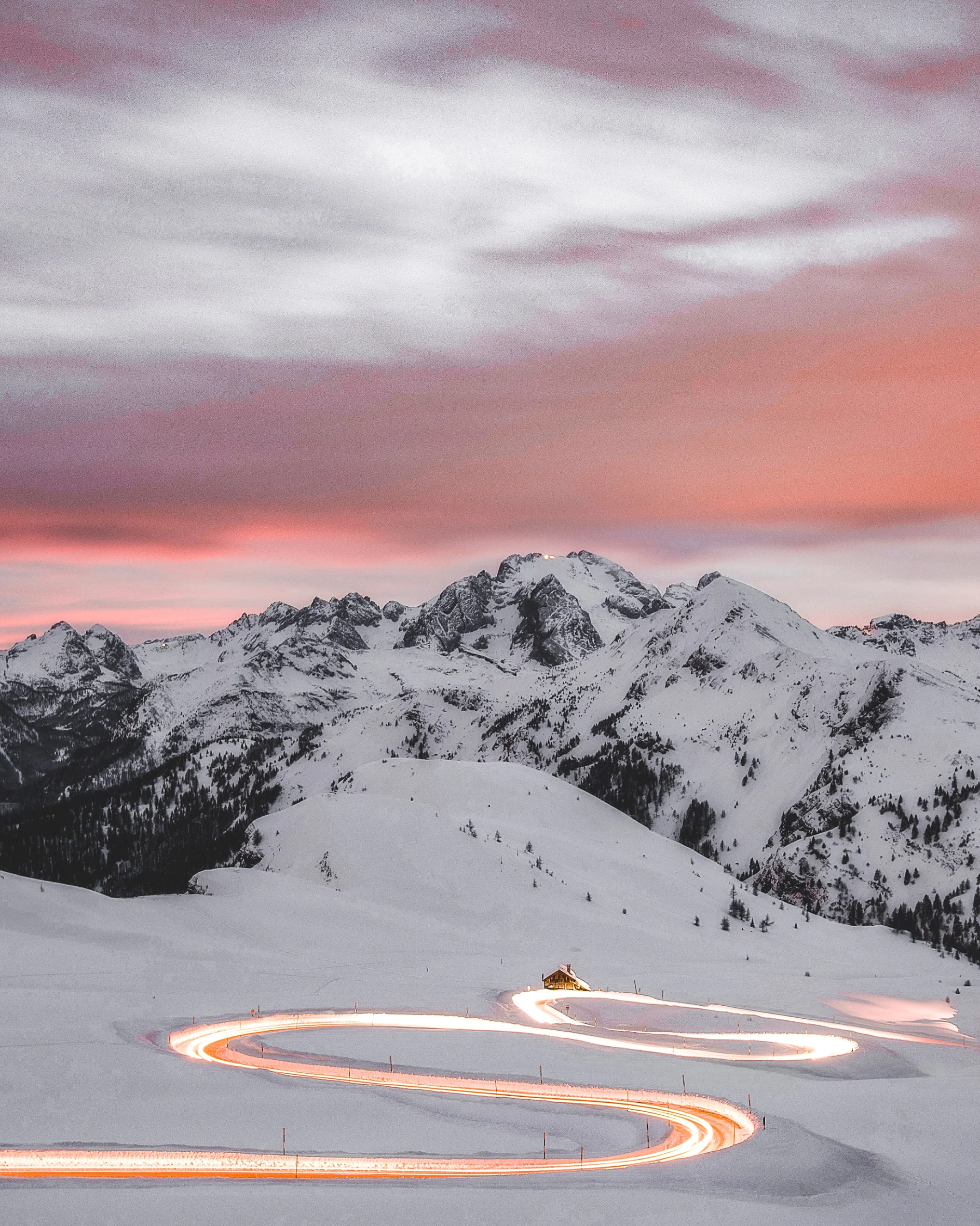 A snow-covered mountain landscape at dusk features a winding road illuminated by car lights. The sky blends pink and gray hues. Near the road, a small building is visible, with mountain peaks in the background. (This text has been generated by AI)