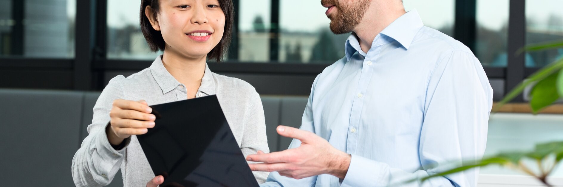 A man and a woman are sitting at a round table, engaged in discussion. The woman holds a tablet, showing it to the man, who gestures with his hand. Papers and tesa tape are on the table. They are in a modern office setting with large windows. (This text has been generated by AI)