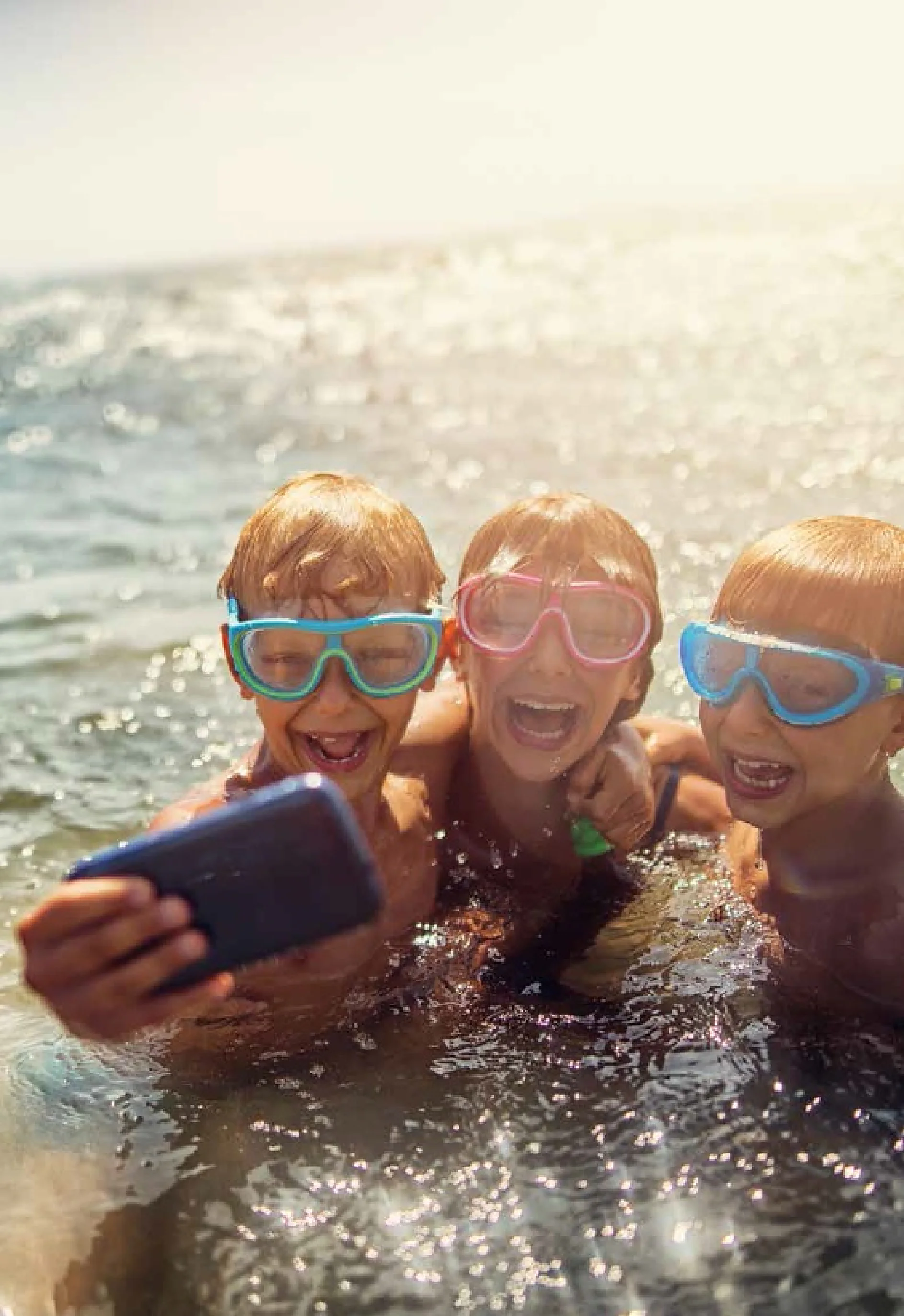 Three children wearing swim goggles smile and pose for a selfie in the water. The sun shines brightly, creating a sparkling effect on the waters surface. One child holds a smartphone to take the photo, while another uses tesa tape to secure their waterproof case. (This text has been generated by AI)