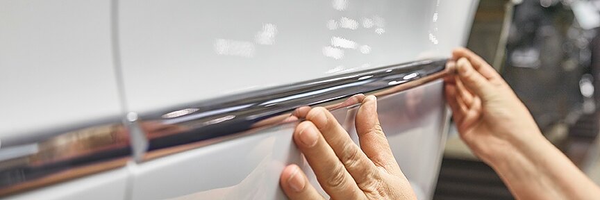 Two hands are applying a chrome trim to the side of a white vehicle using tesa tape. The image shows a close-up of the trim being aligned on the cars body, highlighting the hands and part of the vehicles side panel. (This text has been generated by AI)