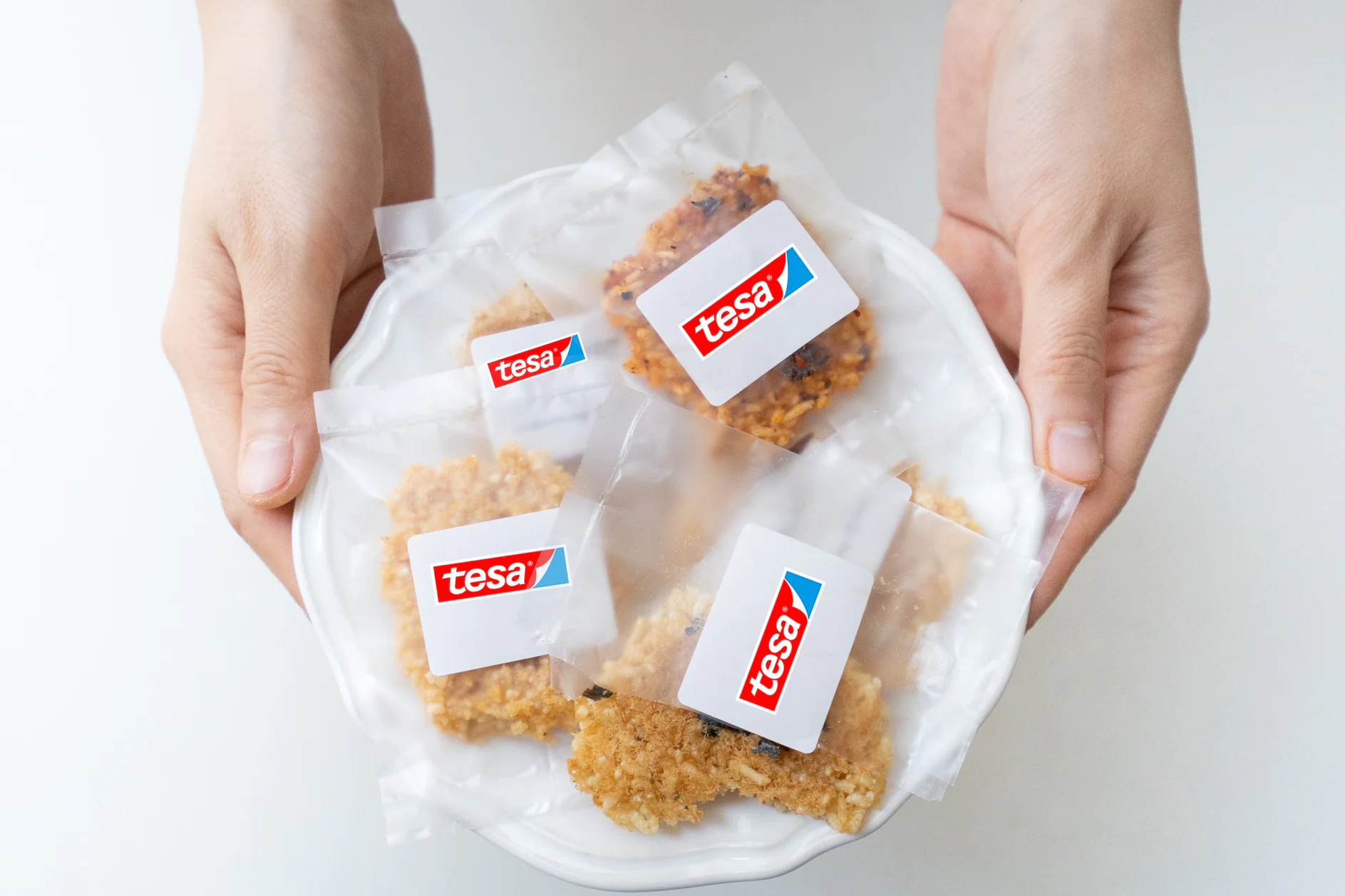 Two hands hold a white plate containing several packaged items. Each package is sealed and features the tesa tape logo, a red and blue design. The packages contain a crumbly, golden-brown substance, possibly food. The background is plain white. (This text has been generated by AI)