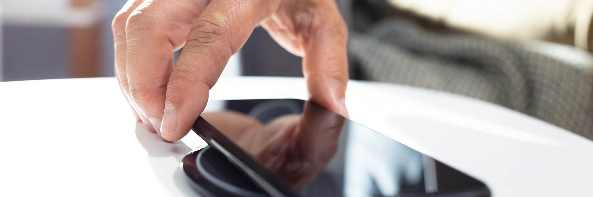 A hand is placing a smartphone on a wireless charging pad on a white table. The smartphone screen is black and reflective. The background is blurred with some furniture visible, and pieces are secured neatly using tesa tape. (This text has been generated by AI)