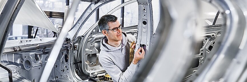 A person in a gray hoodie is inspecting the interior frame of a car in a factory setting. The car structure is metallic and unpainted, showing various parts and components. The environment is bright with large windows in the background. (This text has been generated by AI)