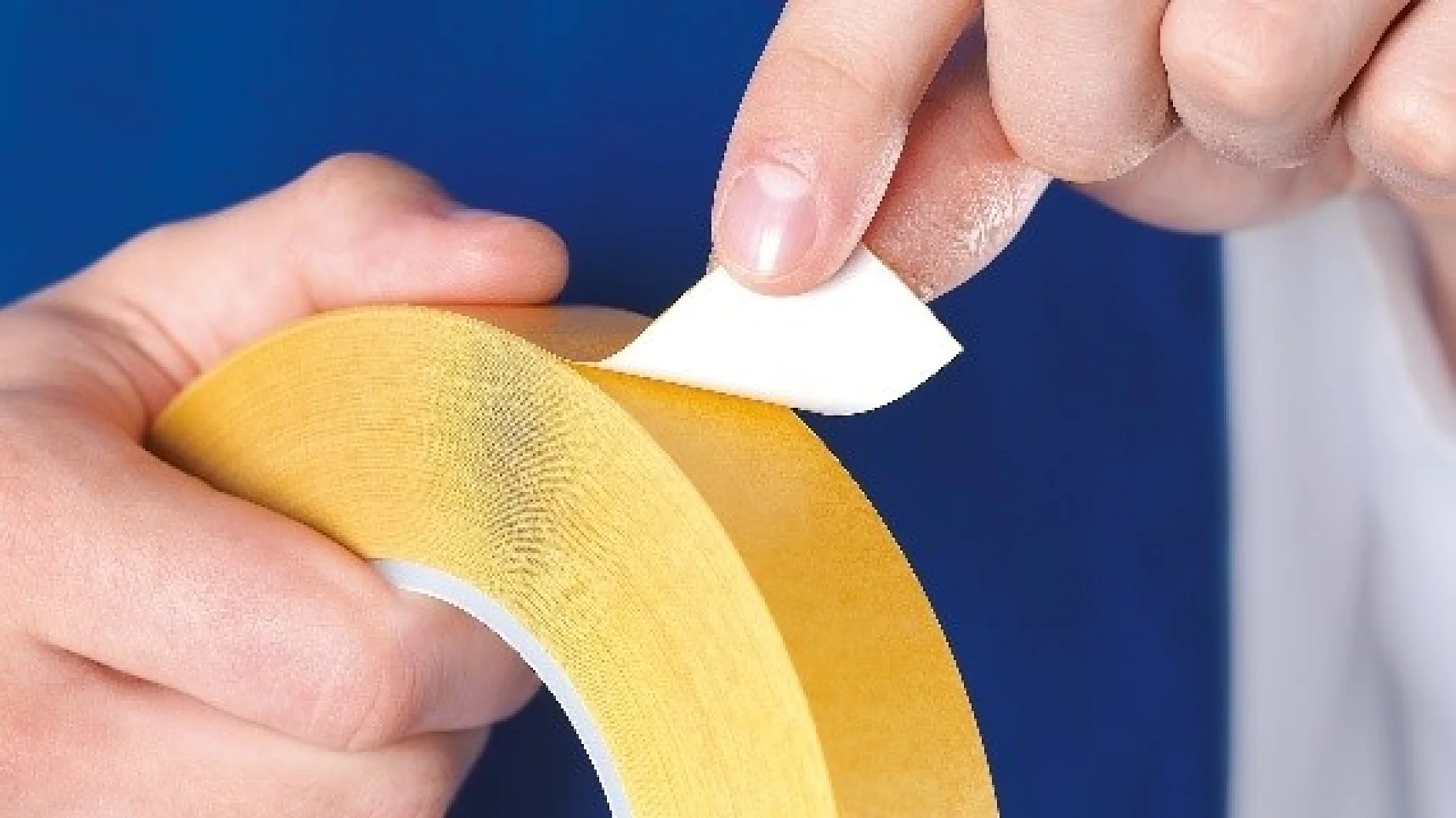 A person is peeling the backing off a roll of double-sided tesa tape. The tesa tape is yellow with a white peel-off layer. The background is blue. (This text has been generated by AI)