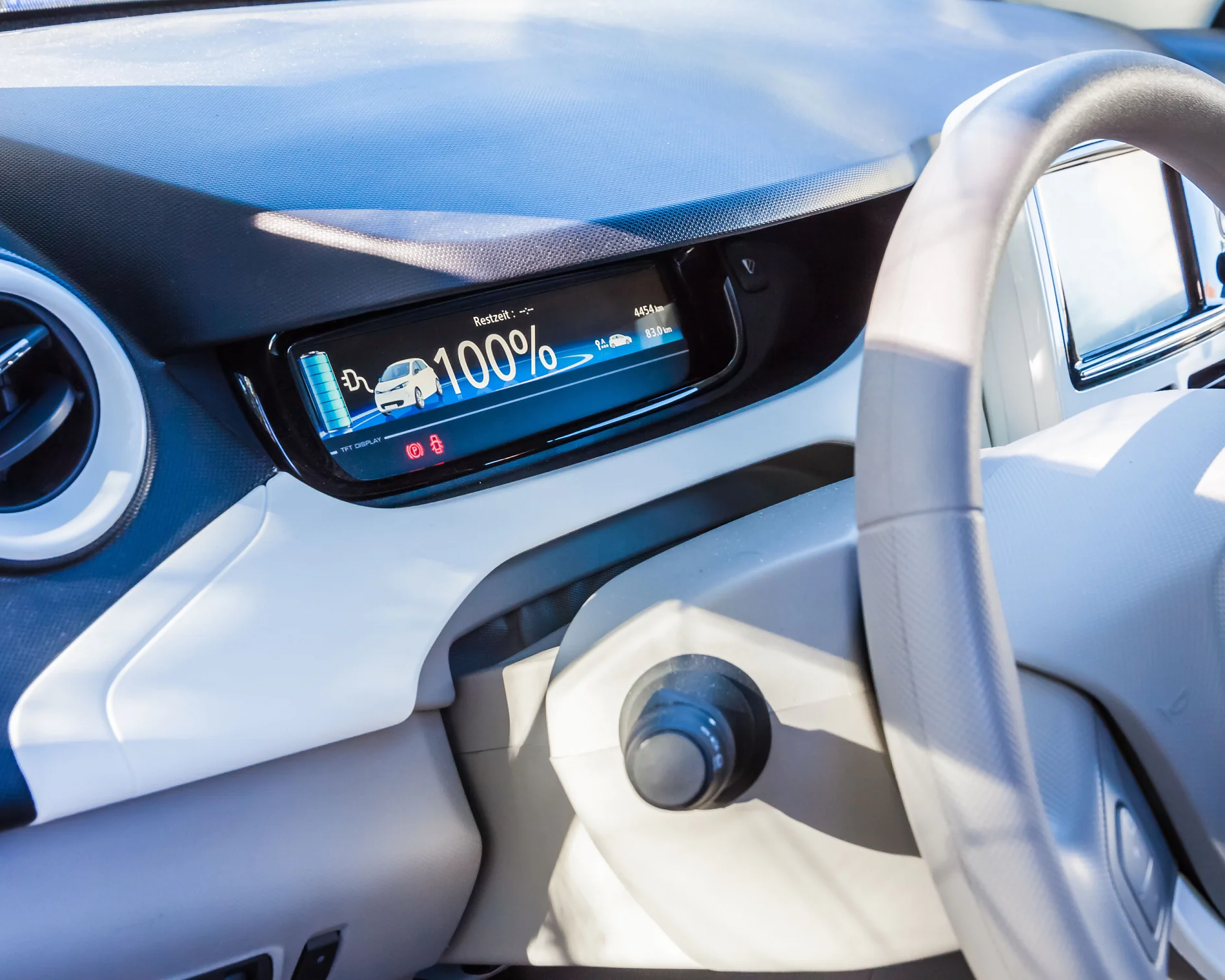 Close-up of a car dashboard featuring a digital screen showing a fully charged electric vehicle battery at 100%. The steering wheel and a round air vent are visible. The interior is light-colored. (This text has been generated by AI)