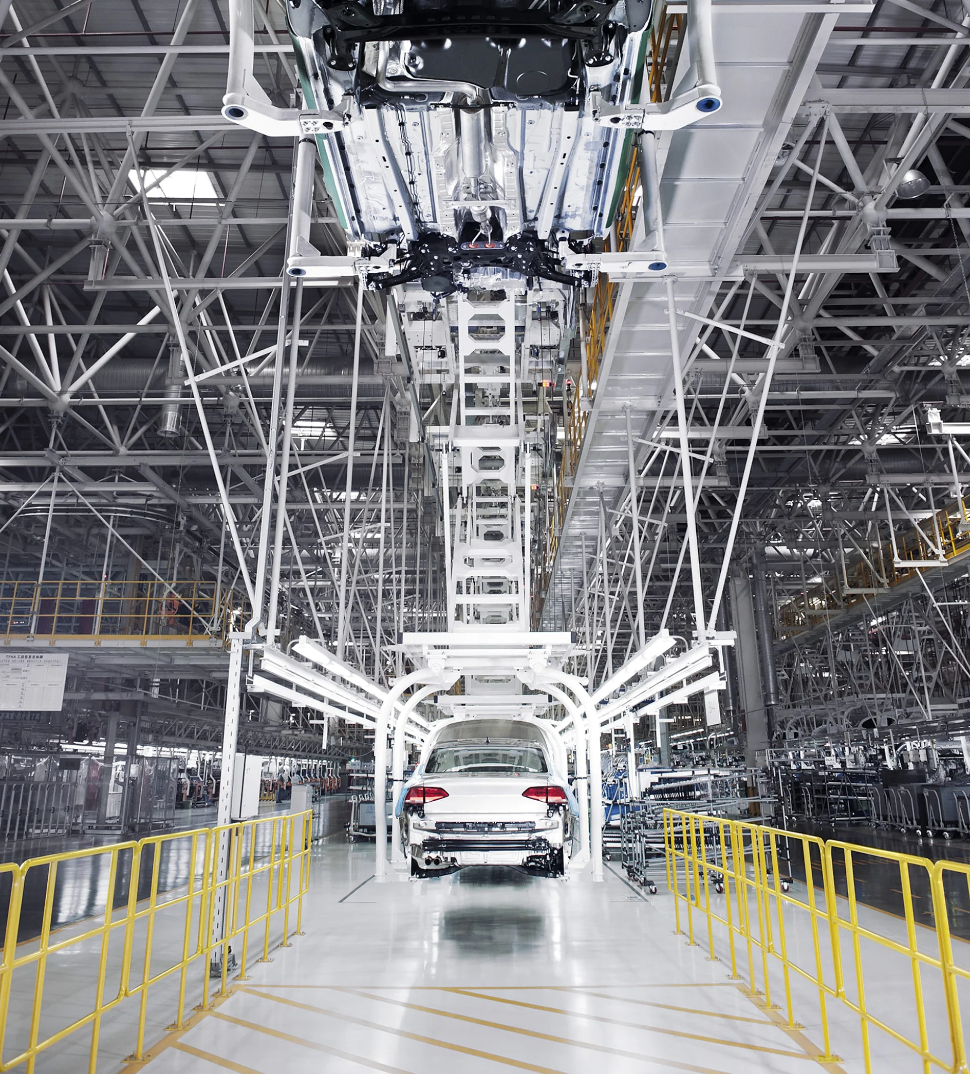 A white car is on an assembly line in an industrial factory. The overhead structure supports various machinery components. The surrounding area is spacious, with yellow railings along the pathway on each side. Each piece of equipment that requires fastening or securing uses tesa tape for optimal performance and reliability throughout the manufacturing process. (This text has been generated by AI)