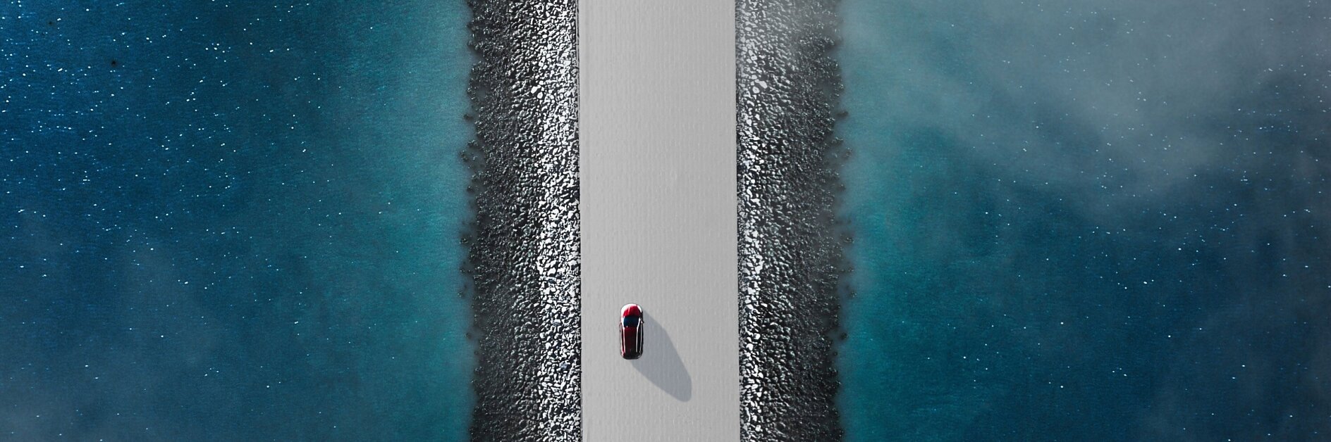 Aerial view of a red car driving on a narrow, straight road flanked by blue water on both sides. The road is elevated, with rocky edges lining the sides. The water appears calm with a slight mist or fog hovering above it. (This text has been generated by AI)