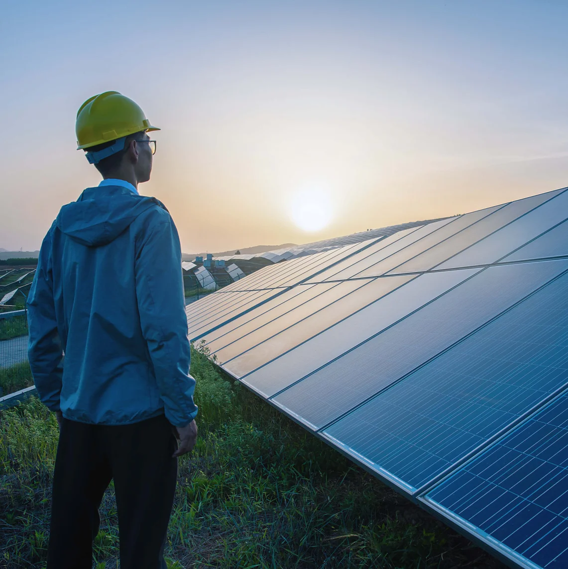 A person in a yellow hard hat and blue jacket stands in a grassy area, looking at a large array of solar panels at sunset. The sky is a gradient of orange and blue, and distant hills are visible under the setting sun. (This text has been generated by AI)