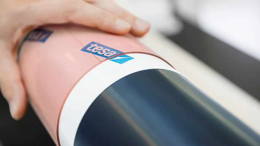 A hand is adjusting a roll of tesa tape. The tesa tape, which is pink and white, is wrapped around a cylindrical object. The background is out of focus, with another hand partially visible. (This text has been generated by AI)