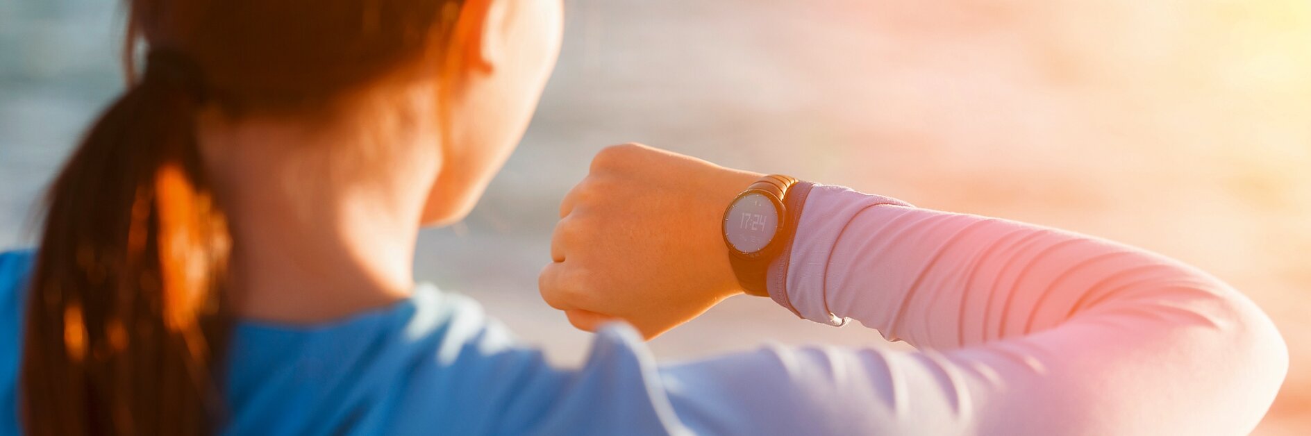 A person with long brown hair is outdoors, looking at a smartwatch on their left wrist. They are wearing a long-sleeve blue top. The background is blurred with sunlight, suggesting a beach or waterfront setting. (This text has been generated by AI)