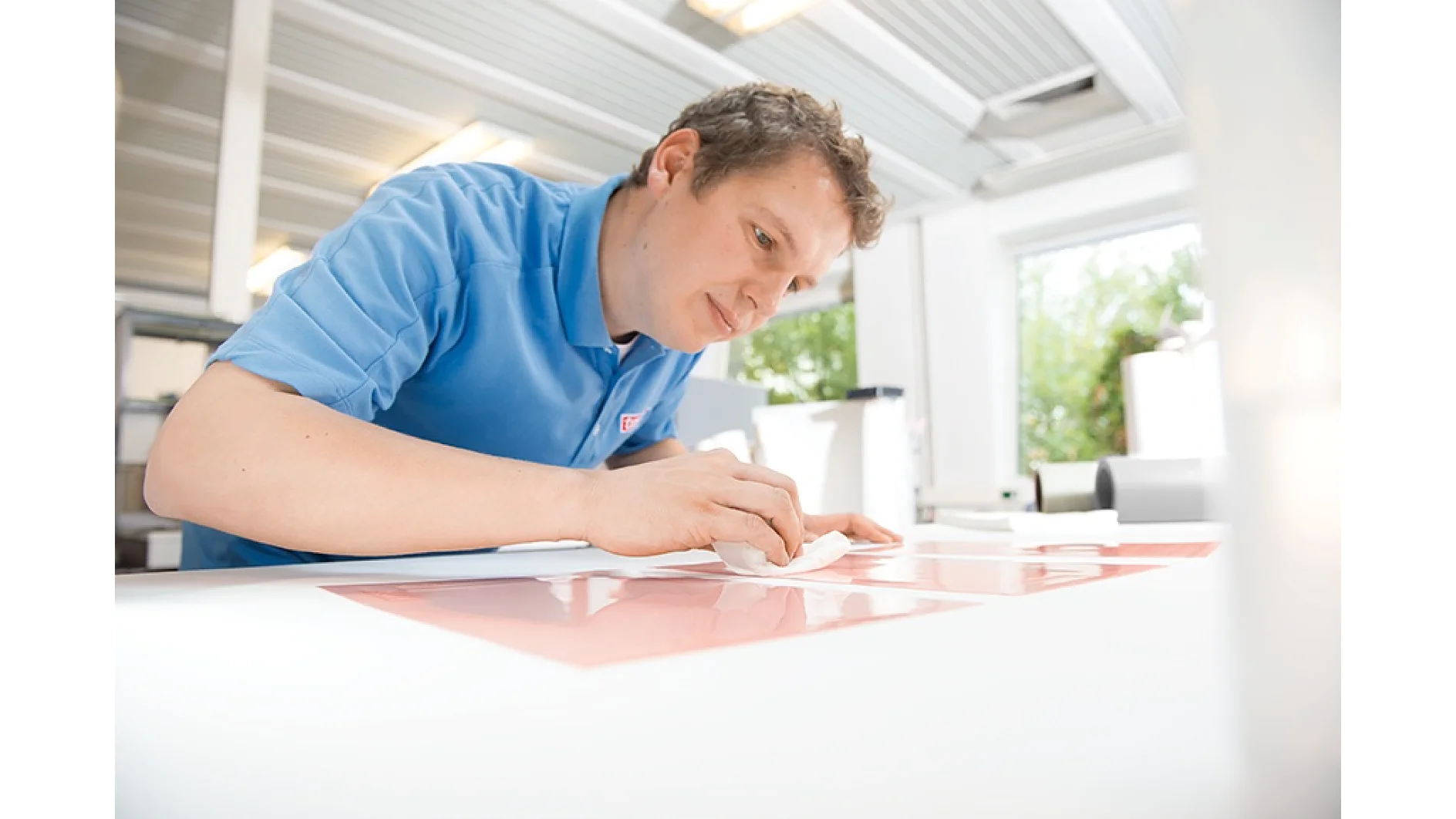 A person in a blue shirt is leaning over a table, focused on applying tesa tape to a surface. The setting appears to be a well-lit workspace with large windows in the background. (This text has been generated by AI)