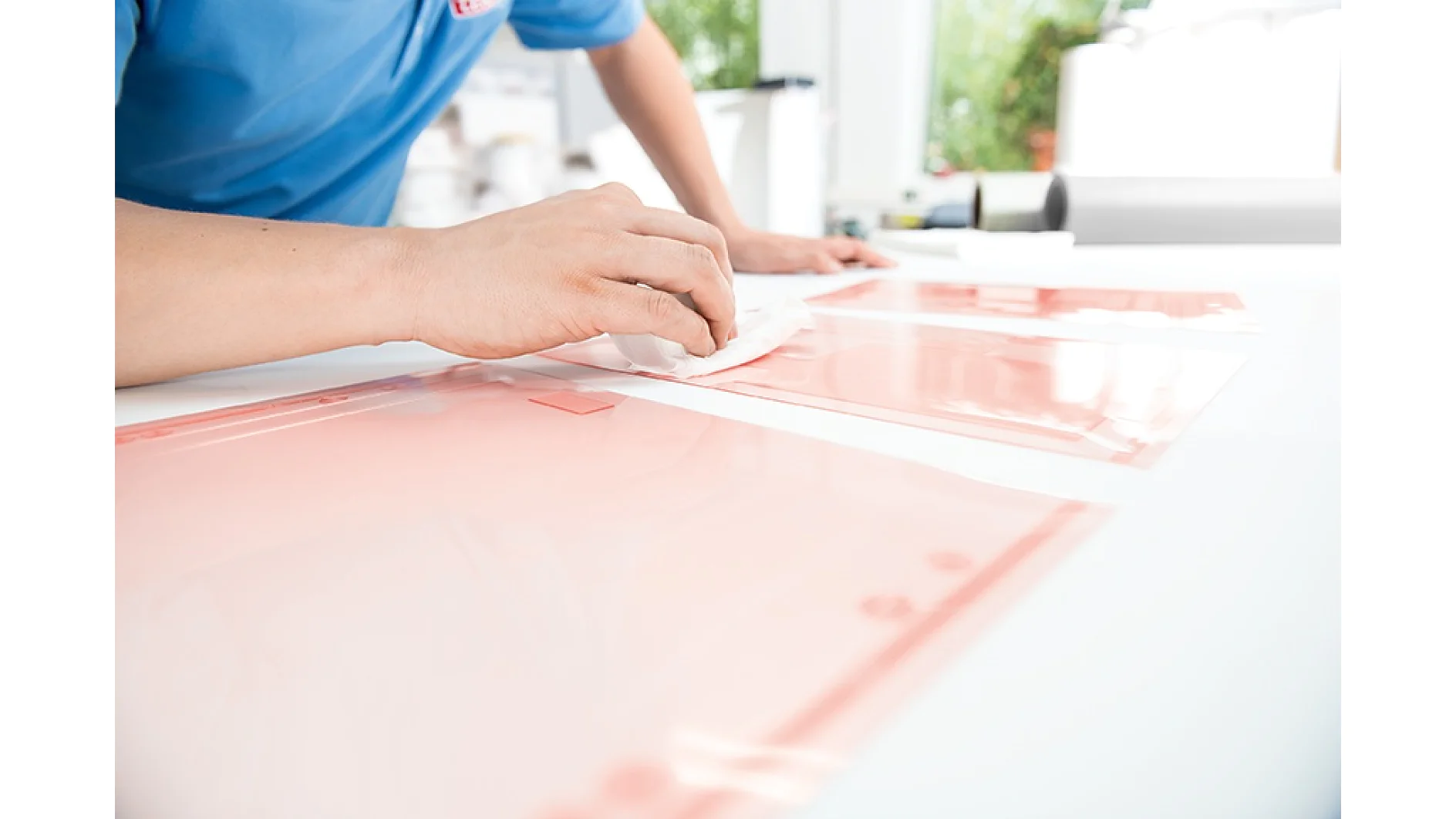 In a blue shirt, a person is wiping a large, clear plastic sheet on a white table with a cloth. The background is bright, showcasing blurred greenery through a window. The focus is on the hand and tesa tape securing the sheet. (This text has been generated by AI)