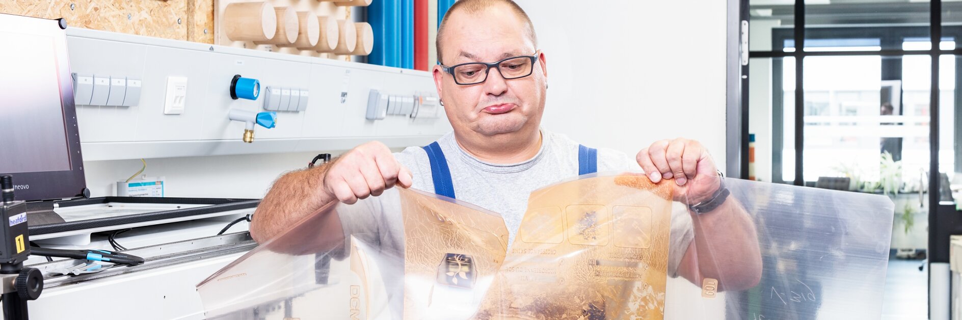 A man wearing glasses and a gray shirt is holding a large, transparent plastic sheet with printed designs secured using tesa tape. He stands in a workroom with machinery, wooden cylinders on shelves, and a visible doorway leading to another area. (This text has been generated by AI)