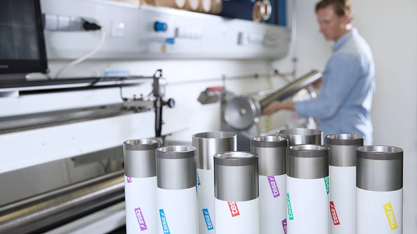 A person in a blue shirt operates machinery in a workshop. In the foreground, there are several cylindrical metal objects with white labels and colored logos. The background includes a computer and various equipment, along with rolls of tesa tape visible on a shelf. (This text has been generated by AI)