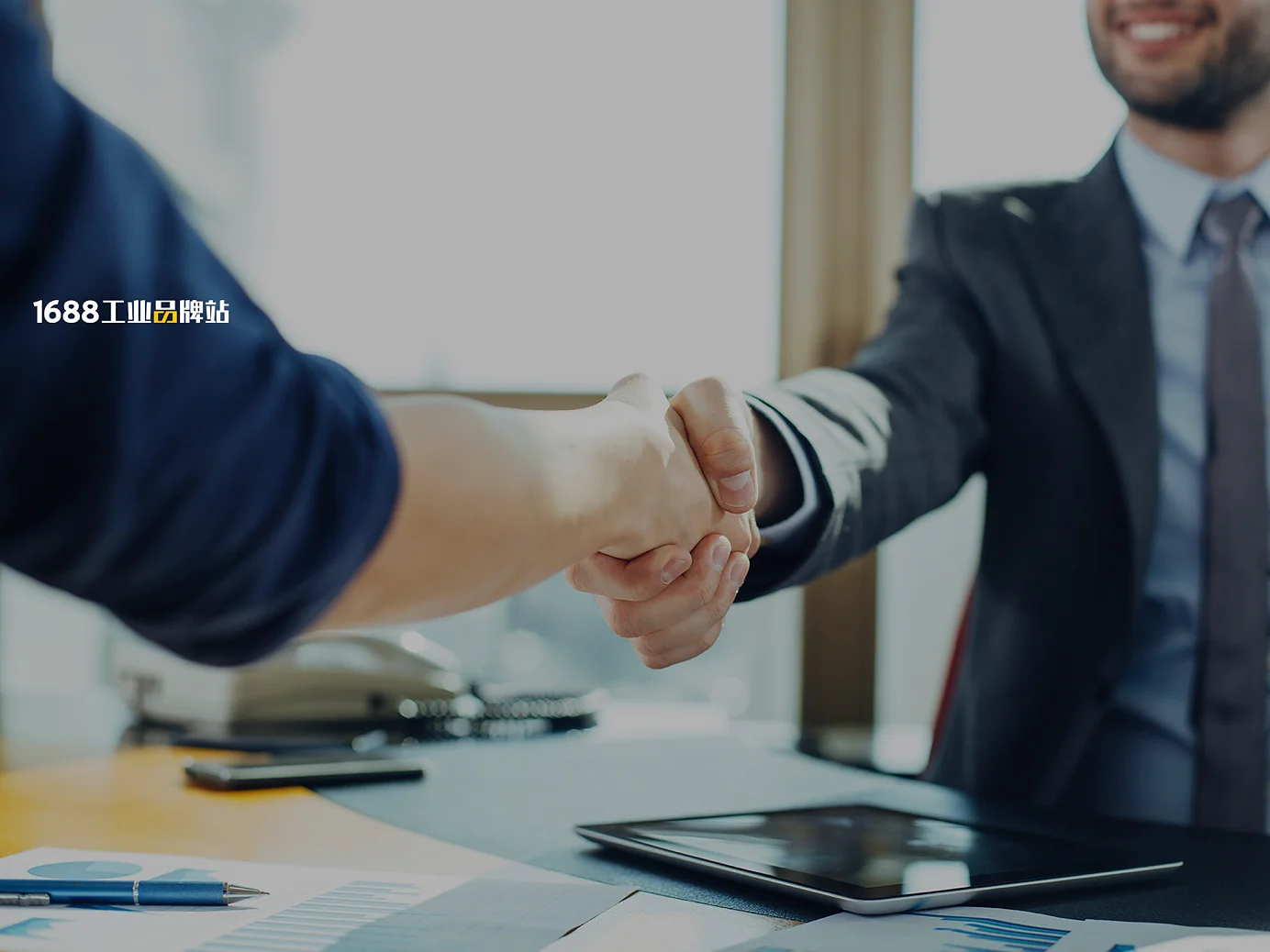 Two people are shaking hands over a desk with documents, charts, and a tablet on it. Both are dressed in business attire. The lighting suggests a daytime office setting. (This text has been generated by AI)