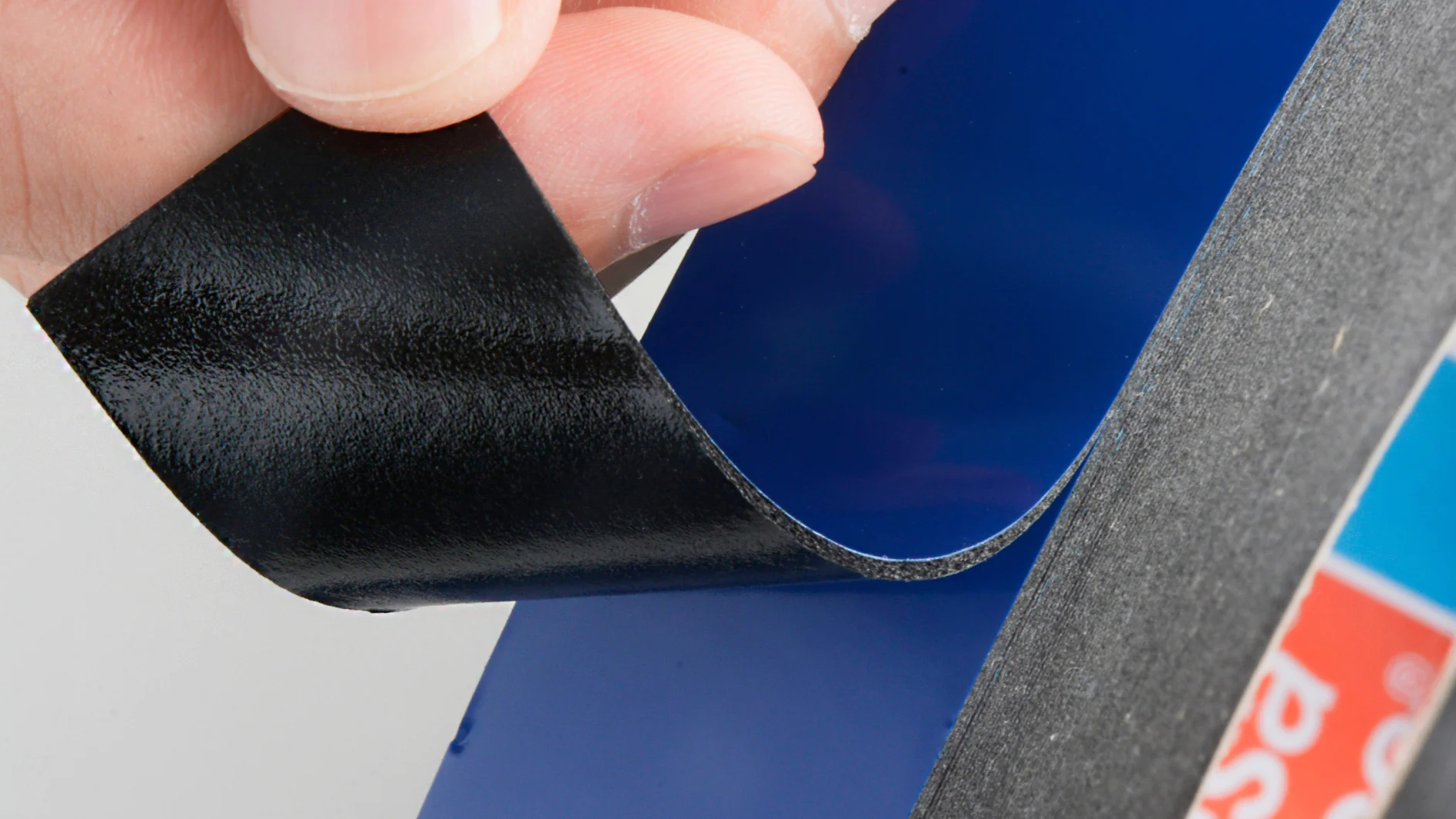 A hand is peeling back the edge of a roll of black tesa tape. The tesa tape has a shiny surface and is partially lifted. The roll includes other colors such as blue, and the side of the roll is visible. (This text has been generated by AI)