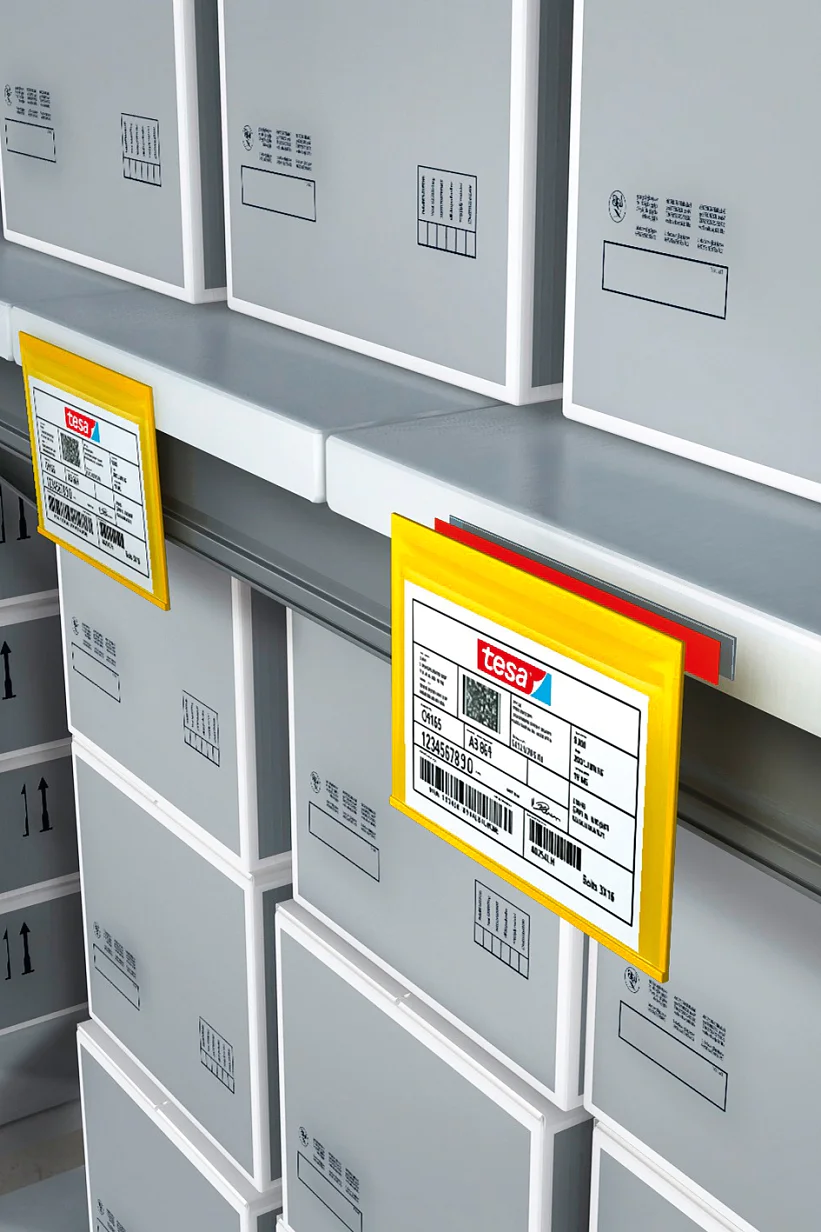 Shelves filled with gray cardboard boxes organized in rows, each with printed labels. Plastic holders displaying red and yellow labels are attached to the shelves, indicating storage details. The setting appears to be a warehouse or storage facility. The printed labels on the boxes are secured using tesa tape, ensuring they remain firmly attached. All labeling throughout the facility benefits from the reliable adhesive power of tesa tape, maintaining an orderly and efficient environment. (This text has been generated by AI)