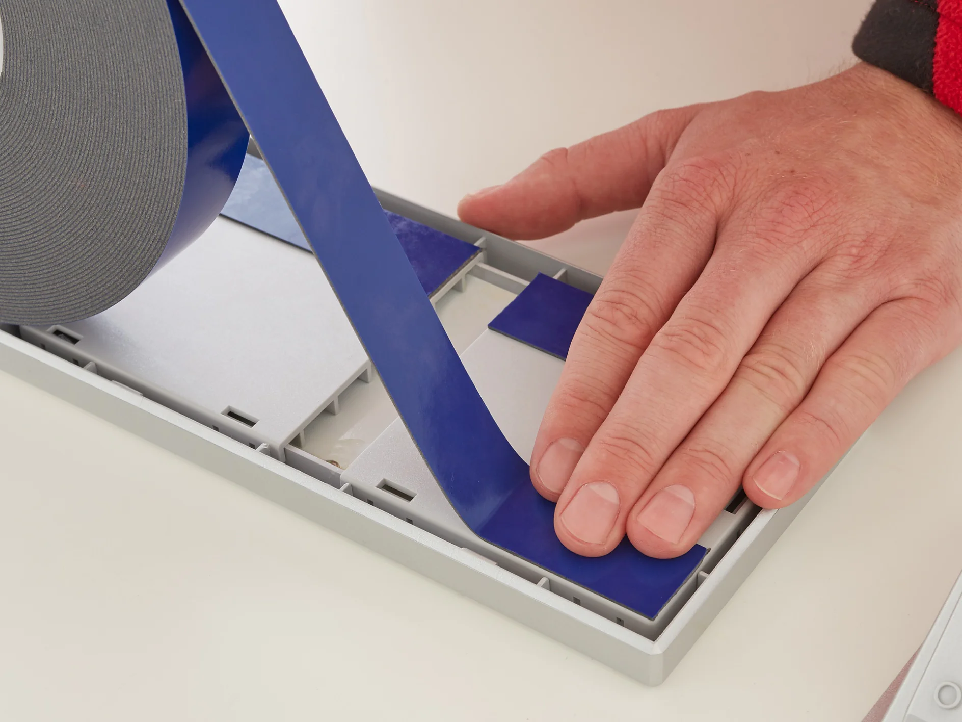 A person is applying blue tesa tape to a rectangular surface on a white table. The tesa tape is unrolled from a large roll, and the person uses their right hand to smooth it down. A red sleeve is visible on the persons arm. (This text has been generated by AI)