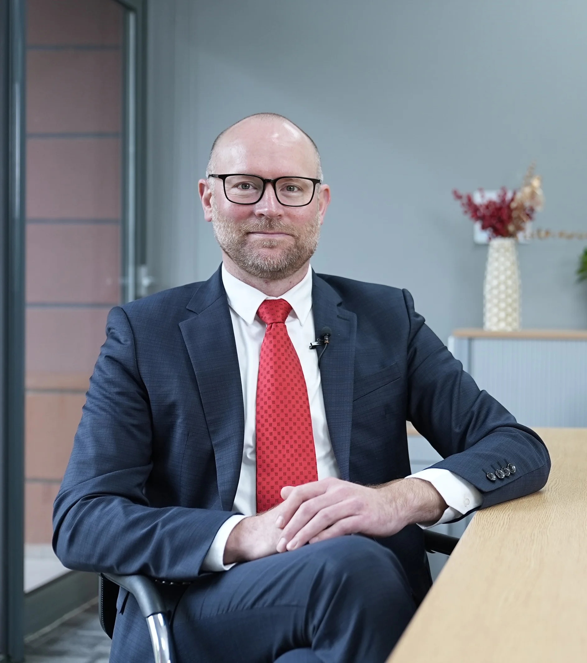 A man with a beard and glasses is seated at a table in an office setting. He is wearing a dark suit, white shirt, and red tie. Behind him are a door and a cabinet with decorative items secured with tesa tape. (This text has been generated by AI)