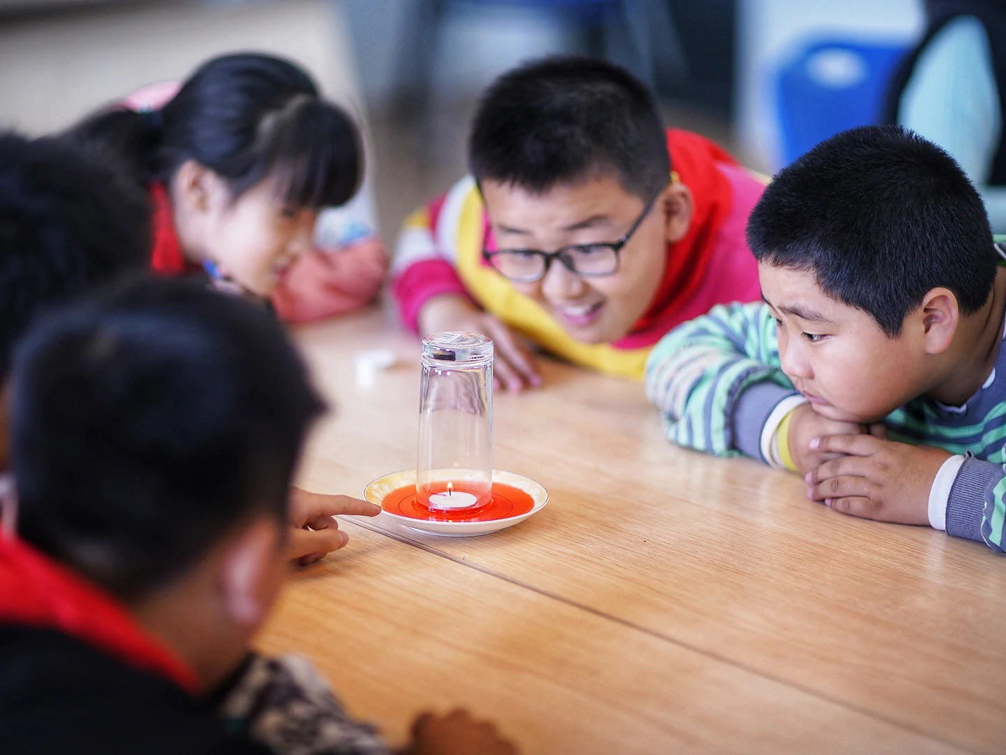 Children are gathered around a table, observing a science experiment. A lit candle is on a plate covered by an inverted glass, secured in place with tesa tape. One child is pointing at it, while the others watch attentively, suggesting they are curious about the experiment. (This text has been generated by AI)