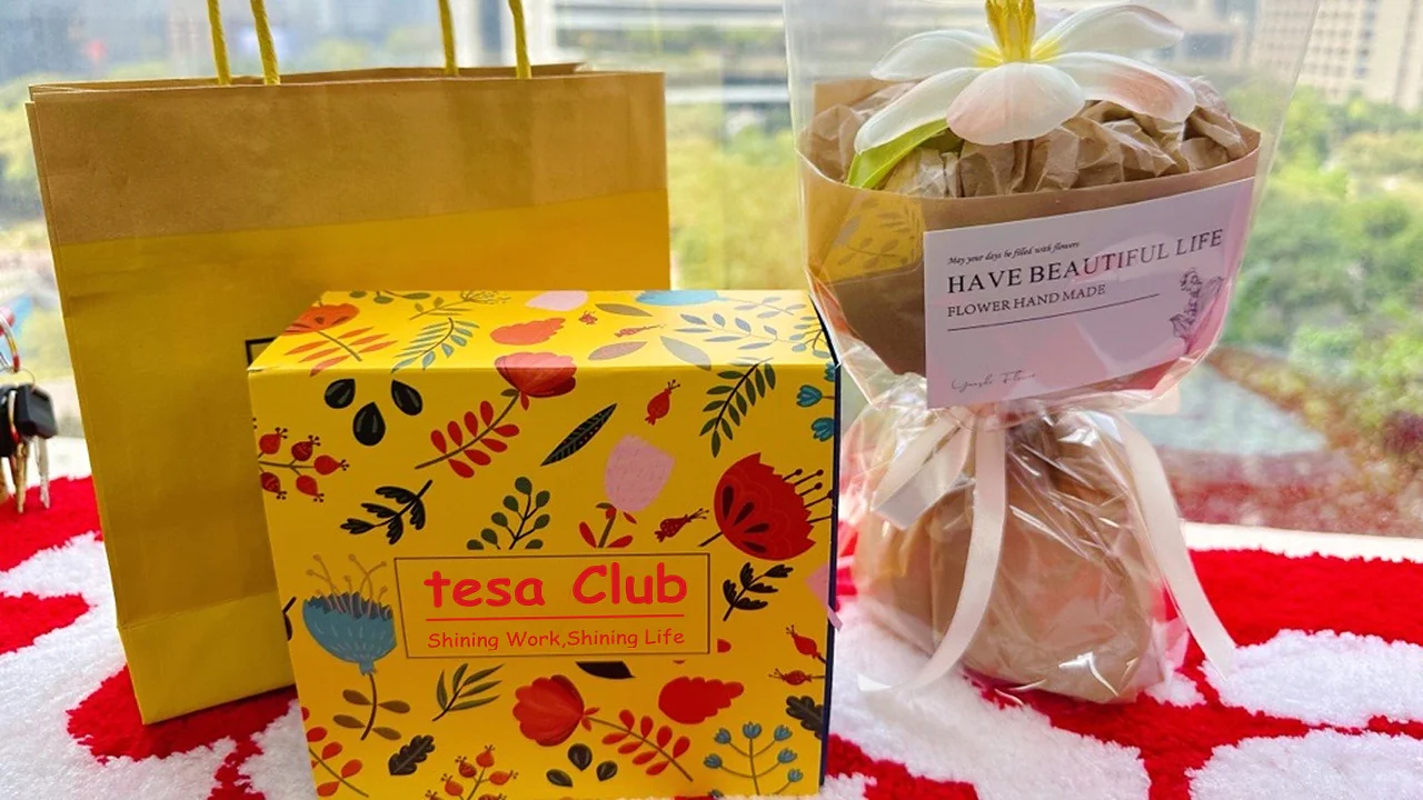 A yellow gift bag is placed next to a bright yellow box with colorful floral patterns. The box says tesa Club. On the right, there is a wrapped gift held together with tesa tape, featuring a label HAVE BEAUTIFUL LIFE and a flower decoration on top. (This text has been generated by AI)