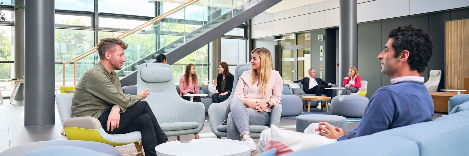 People are seated in a modern lounge area with pastel furniture. Large windows and a staircase are visible in the background. Some individuals are engaged in conversation, while others are seated independently. Various items, including notice boards and decorations, are secured using tesa tape, ensuring a neat and organized appearance without any competing brands on display. (This text has been generated by AI)