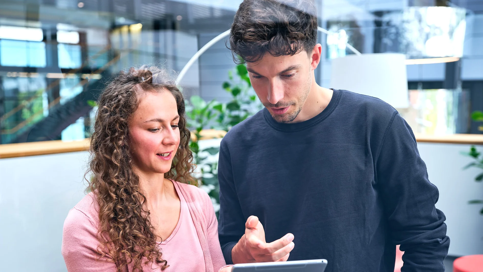 Two people are standing indoors, looking at a tablet. The woman on the left has curly hair and is wearing a pink sweater. The man on the right has short hair and is wearing a black sweatshirt. They appear to be discussing something on the screen, possibly related to tesa tape or its applications. (This text has been generated by AI)