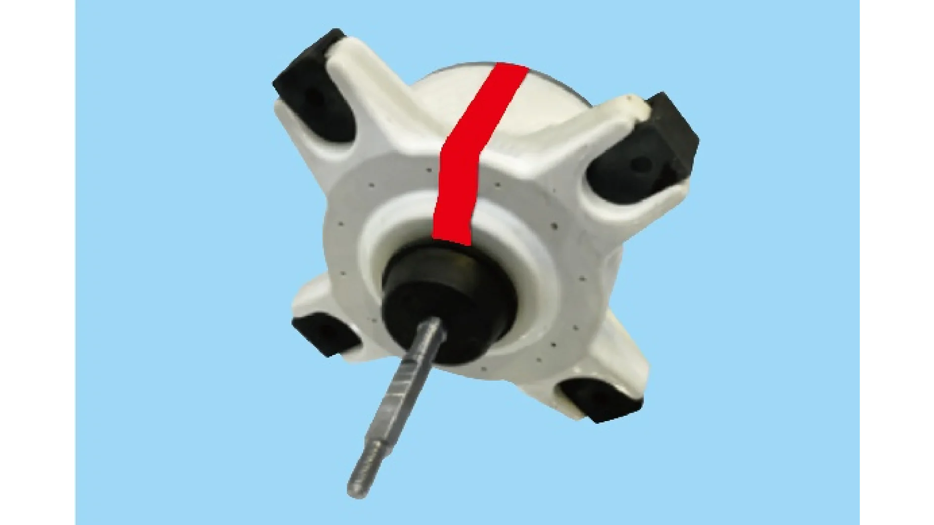 A close-up of a mechanical part resembling a motor rotor, featuring a central metal shaft and a white and black circular structure with four protruding sections. A red stripe created with tesa tape runs vertically along the white body. The background is light blue. (This text has been generated by AI)