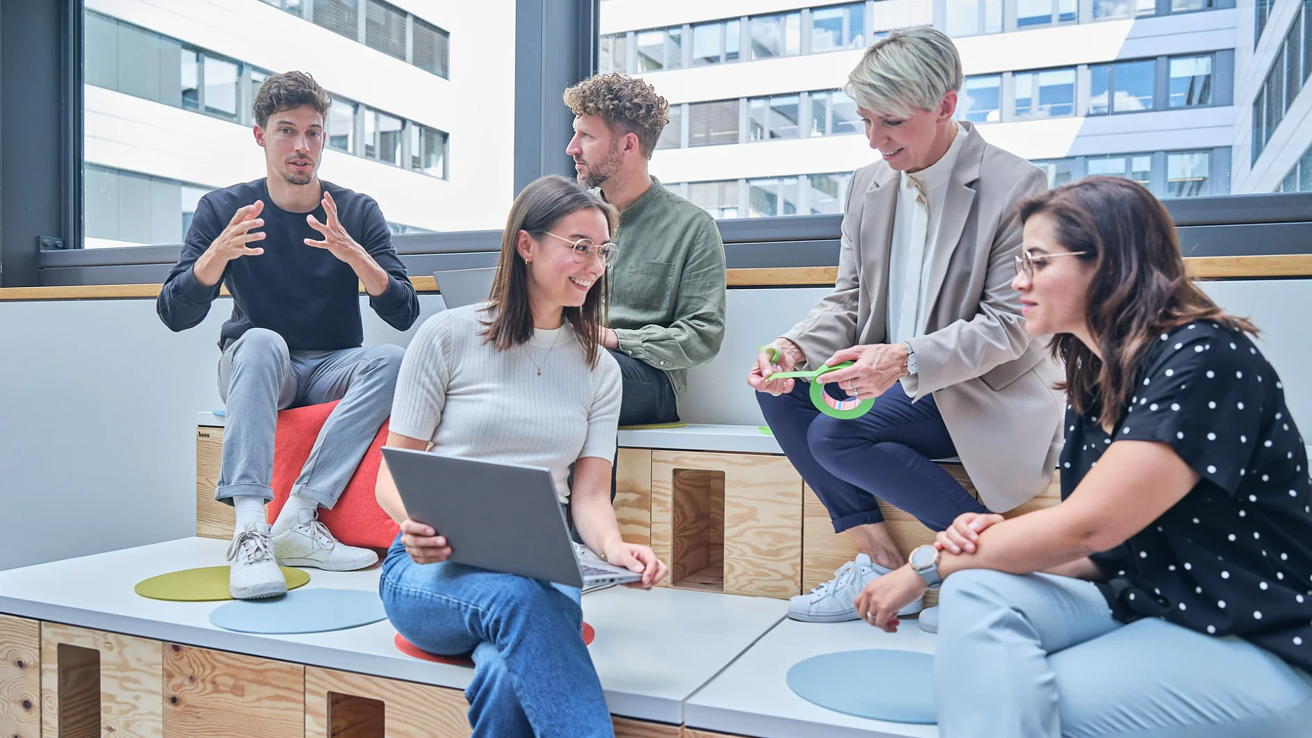 Five people are sitting on tiered wooden seating in a modern office, engaged in discussion. One person is gesturing with hands, another is holding a laptop, and another has a roll of tesa tape. Large windows reveal an urban backdrop. (This text has been generated by AI)