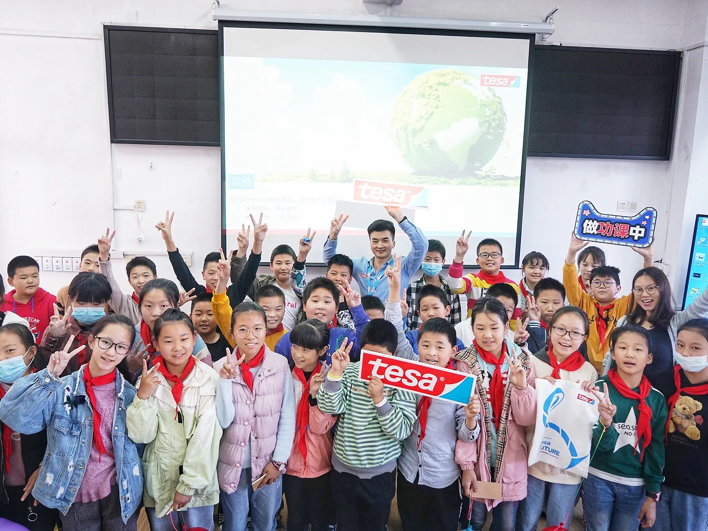 A group of children and a few adults stand in a classroom, smiling and posing with peace signs. Some children hold signs with the word tesa. A projection screen in the background displays an image of a globe and sky. (This text has been generated by AI)