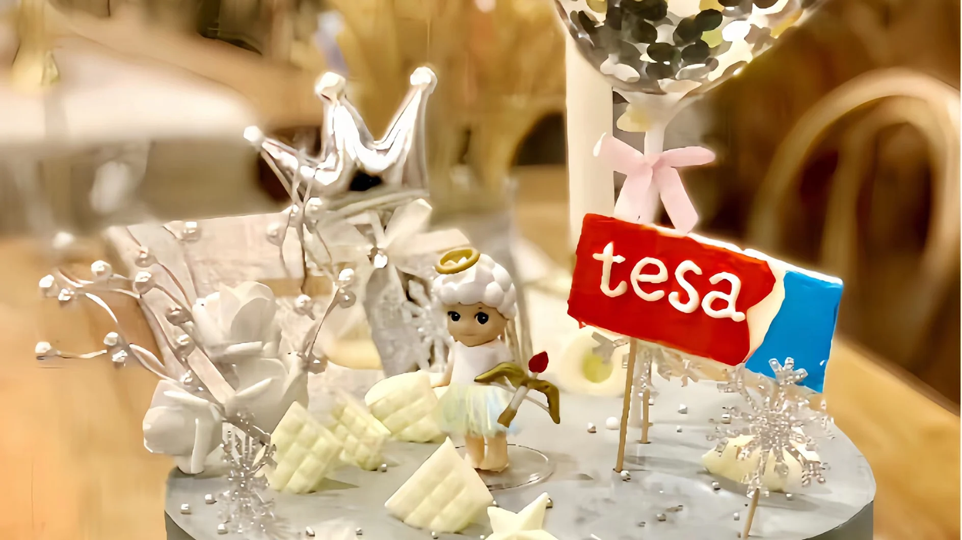 A decorative cake with a small angel figurine, white flowers, and silver accents. A red and blue sign reading tesa is placed on top. A large balloon with a dark and light pattern hovers above. (This text has been generated by AI)