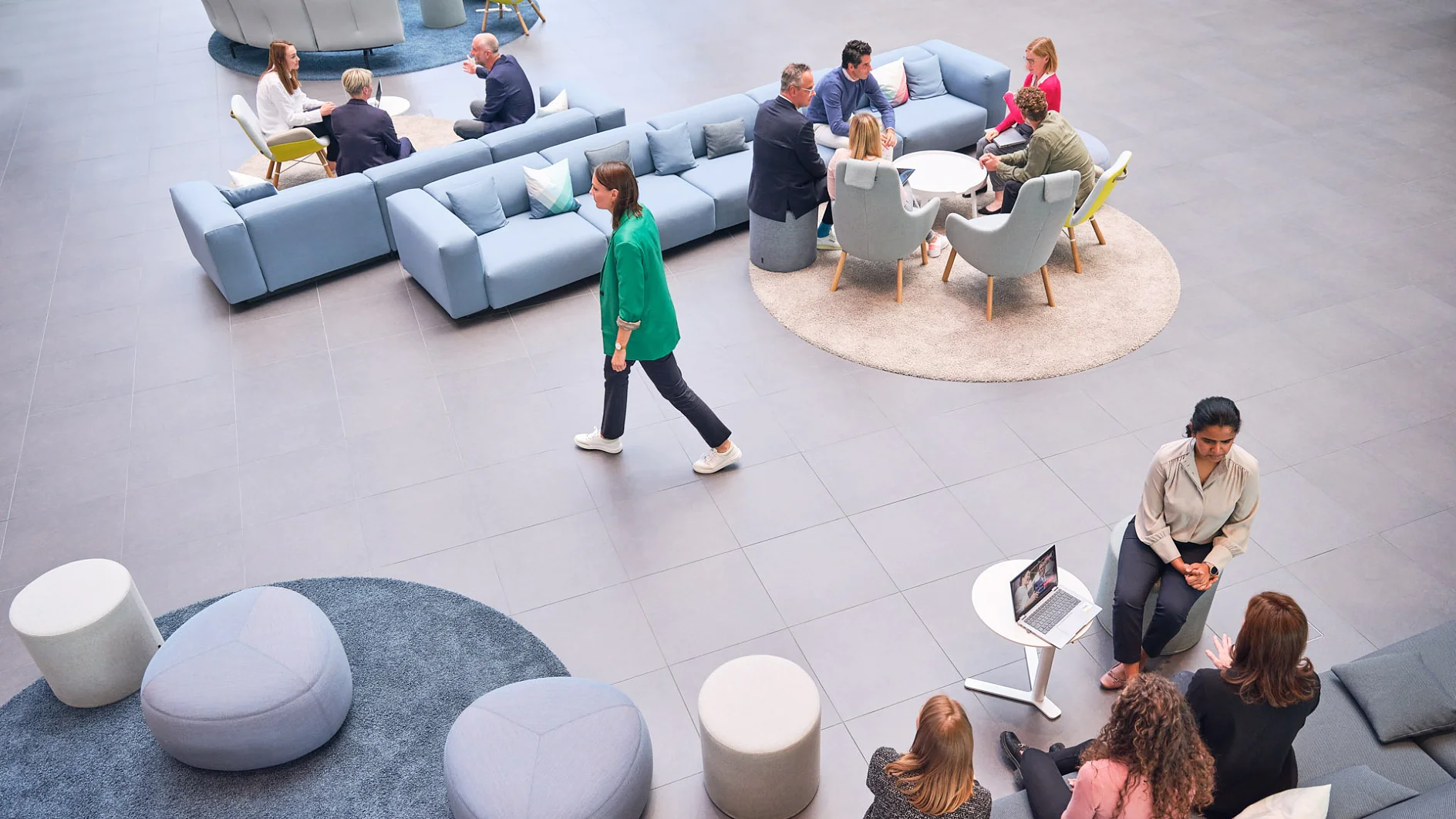 A spacious, modern office lounge features people sitting on sofas and chairs around tables. One person walks through the area, while a laptop is positioned on a small round table. Light colors and minimalist furniture contribute to a casual atmosphere. (This text has been generated by AI)