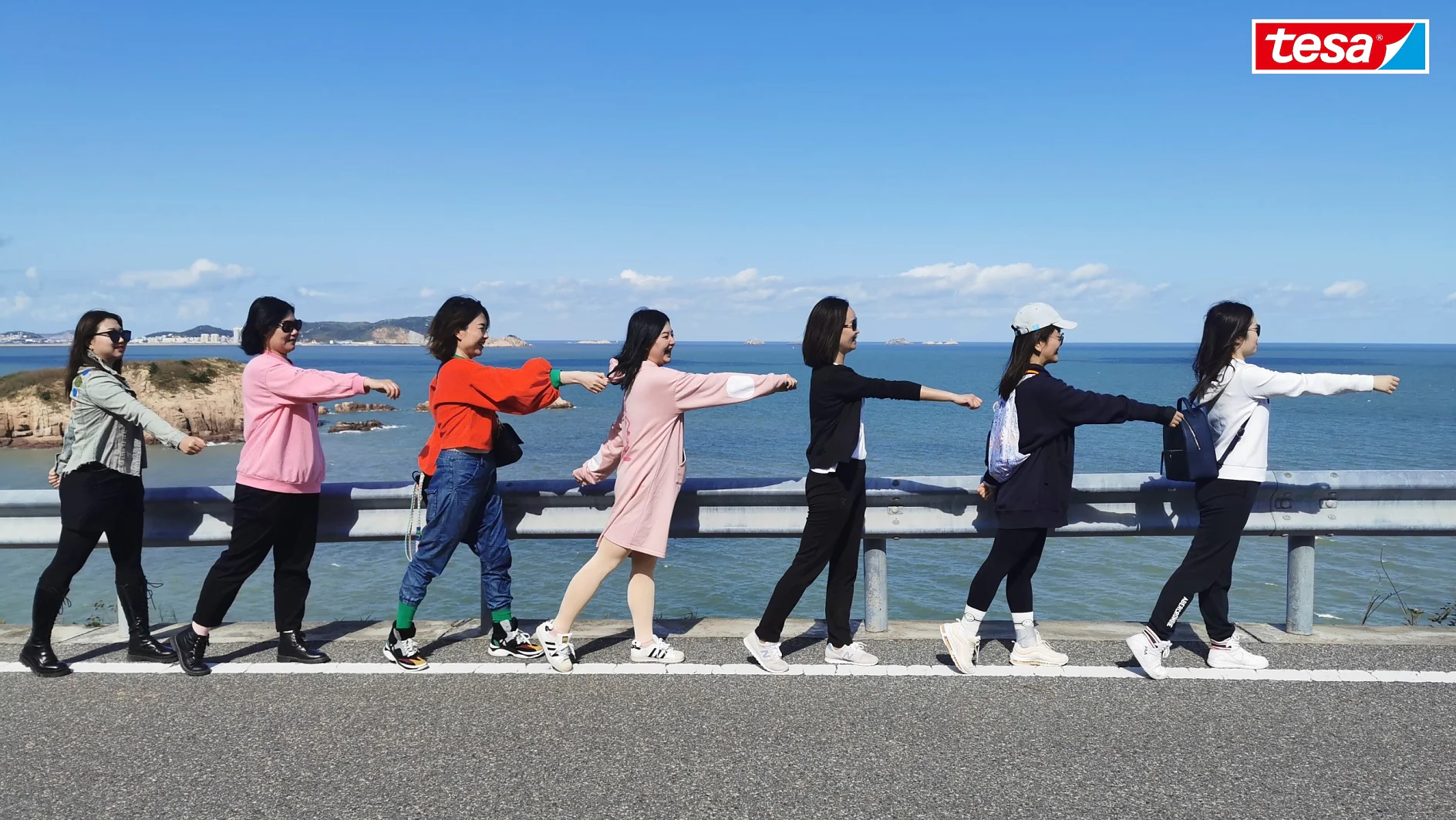 Seven people dressed in casual clothing walk in single file along a road beside a body of water, their arms outstretched. The sky is clear and blue. A tesa tape logo is in the top right corner of the image. (This text has been generated by AI)