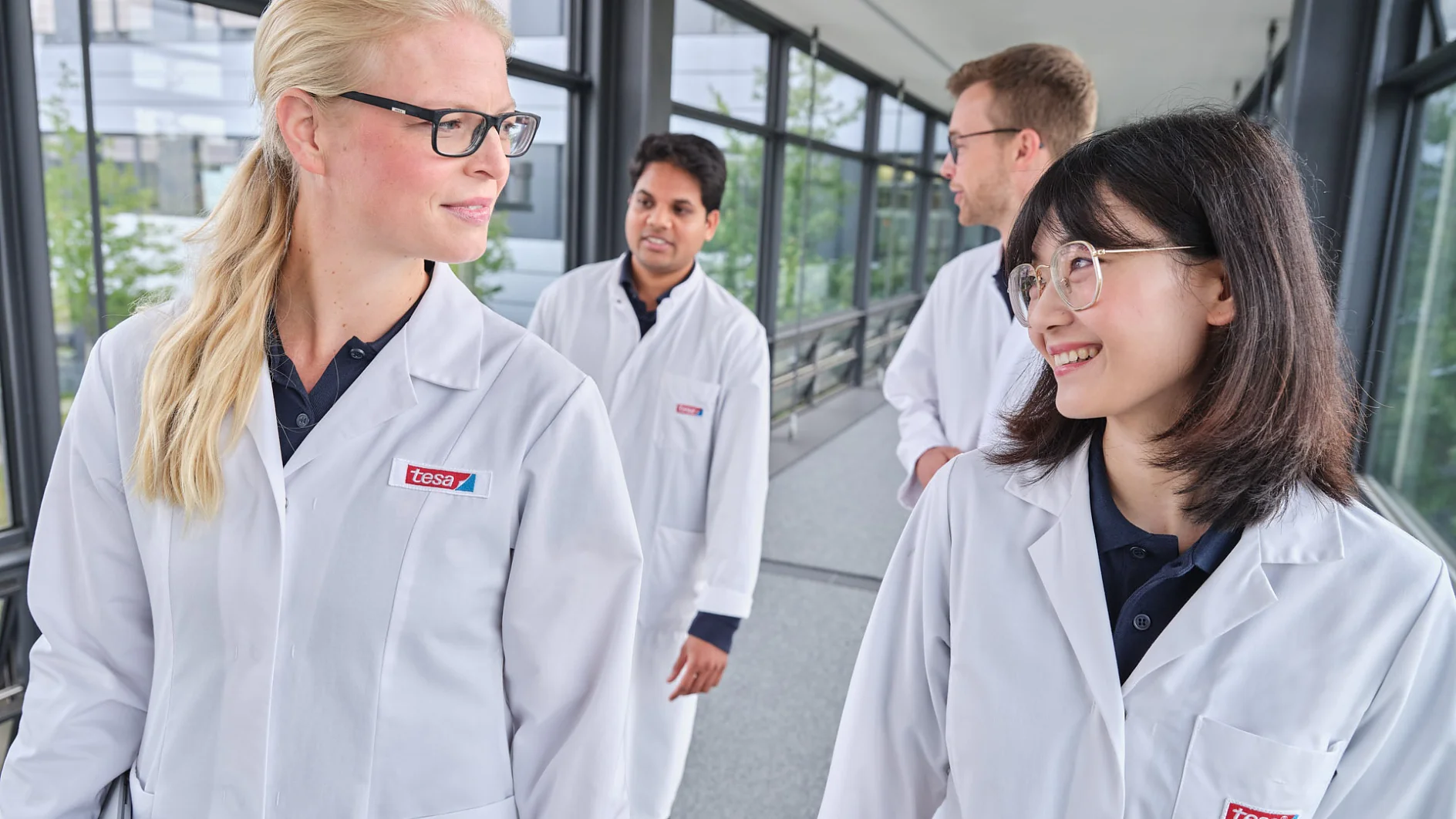 Four people in lab coats walk together along a corridor with large windows. Two individuals in the foreground, one with glasses and long blonde hair, and another with glasses and long dark hair, are looking at each other and smiling. (This text has been generated by AI)