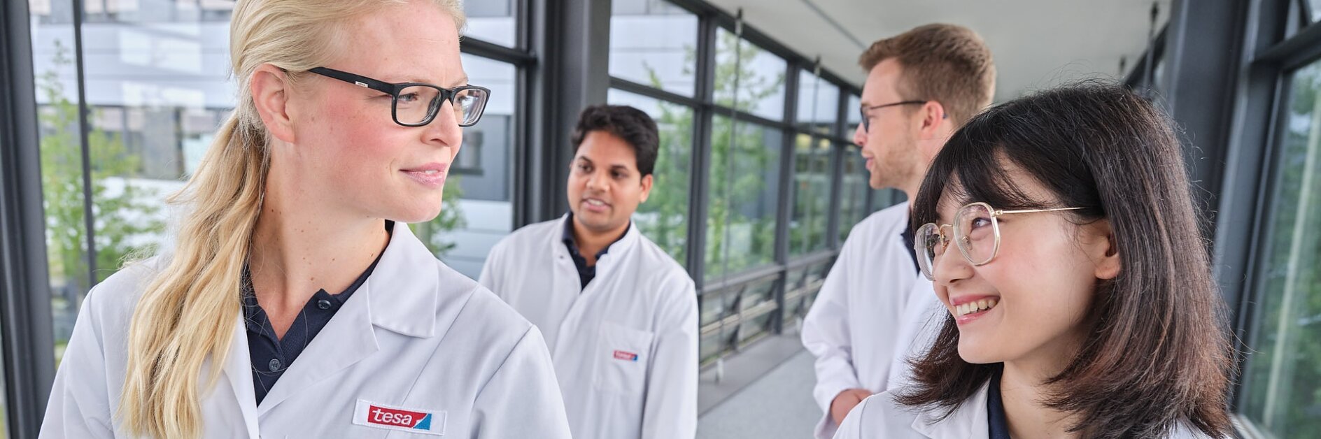 Four people in lab coats walk together along a corridor with large windows. Two individuals in the foreground, one with glasses and long blonde hair, and another with glasses and long dark hair, are looking at each other and smiling. (This text has been generated by AI)