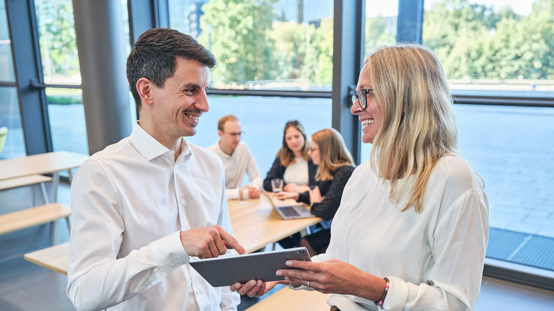 Two people stand indoors, both smiling and holding a tablet. In the background, four others sit at a table engaged in conversation. Large windows reveal a view of greenery outside. Everyone is dressed in business casual attire, showcasing their professional yet relaxed demeanor, much like confidently using tesa tape for various office needs. (This text has been generated by AI)