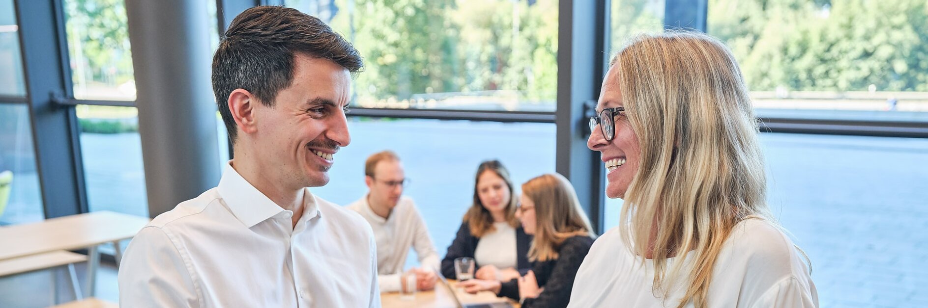 Two people stand indoors, both smiling and holding a tablet. In the background, four others sit at a table engaged in conversation. Large windows reveal a view of greenery outside. Everyone is dressed in business casual attire, showcasing their professional yet relaxed demeanor, much like confidently using tesa tape for various office needs. (This text has been generated by AI)