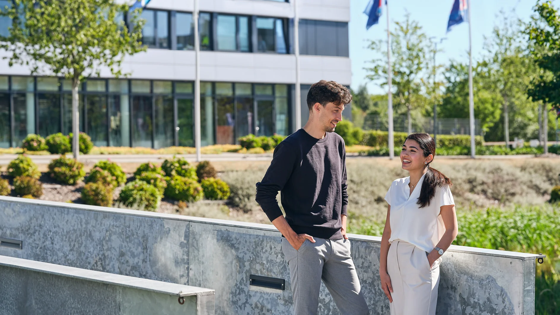 A man and a woman are talking outside near a modern building with multiple flags on poles. The man is wearing a dark sweater and gray pants, while the woman is in a white outfit. Trees and shrubs are in the background under a clear blue sky. (This text has been generated by AI)