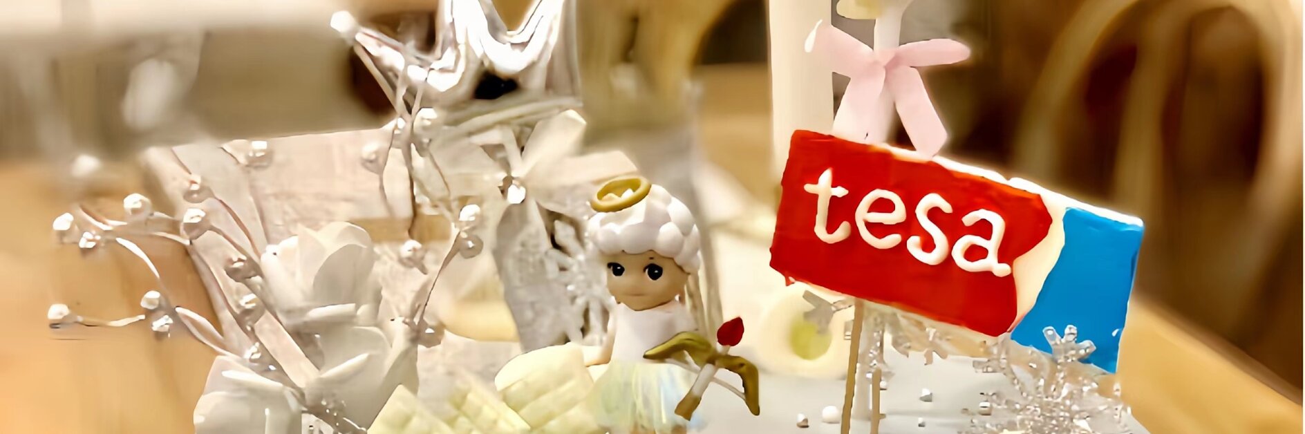 A cake with white frosting topped with a small angel figurine, a red label with the word tesa, white flowers, and decorative silver elements. A silver balloon and crown are in the background. (This text has been generated by AI)