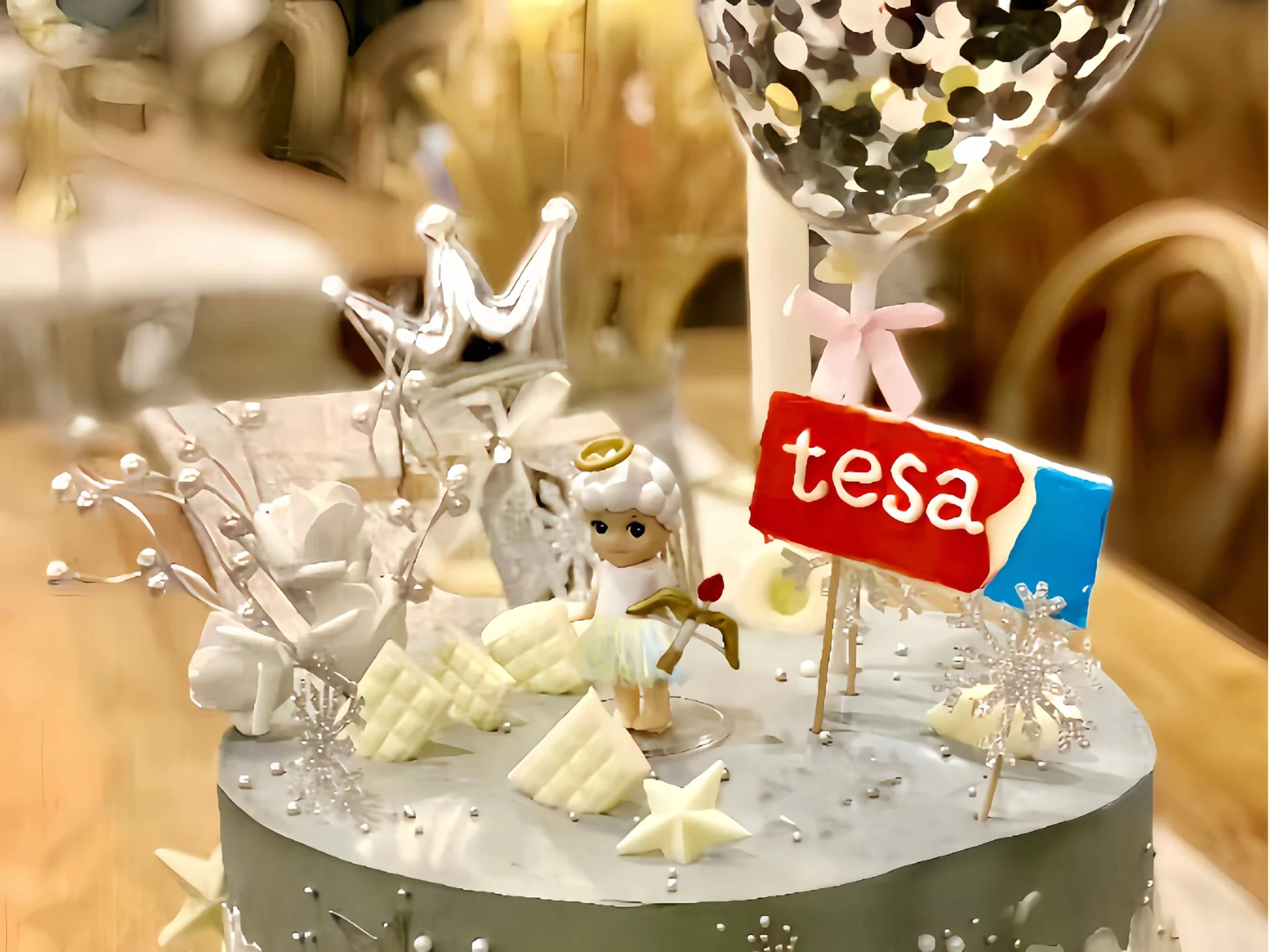 A cake with white frosting topped with a small angel figurine, a red label with the word tesa, white flowers, and decorative silver elements. A silver balloon and crown are in the background. (This text has been generated by AI)